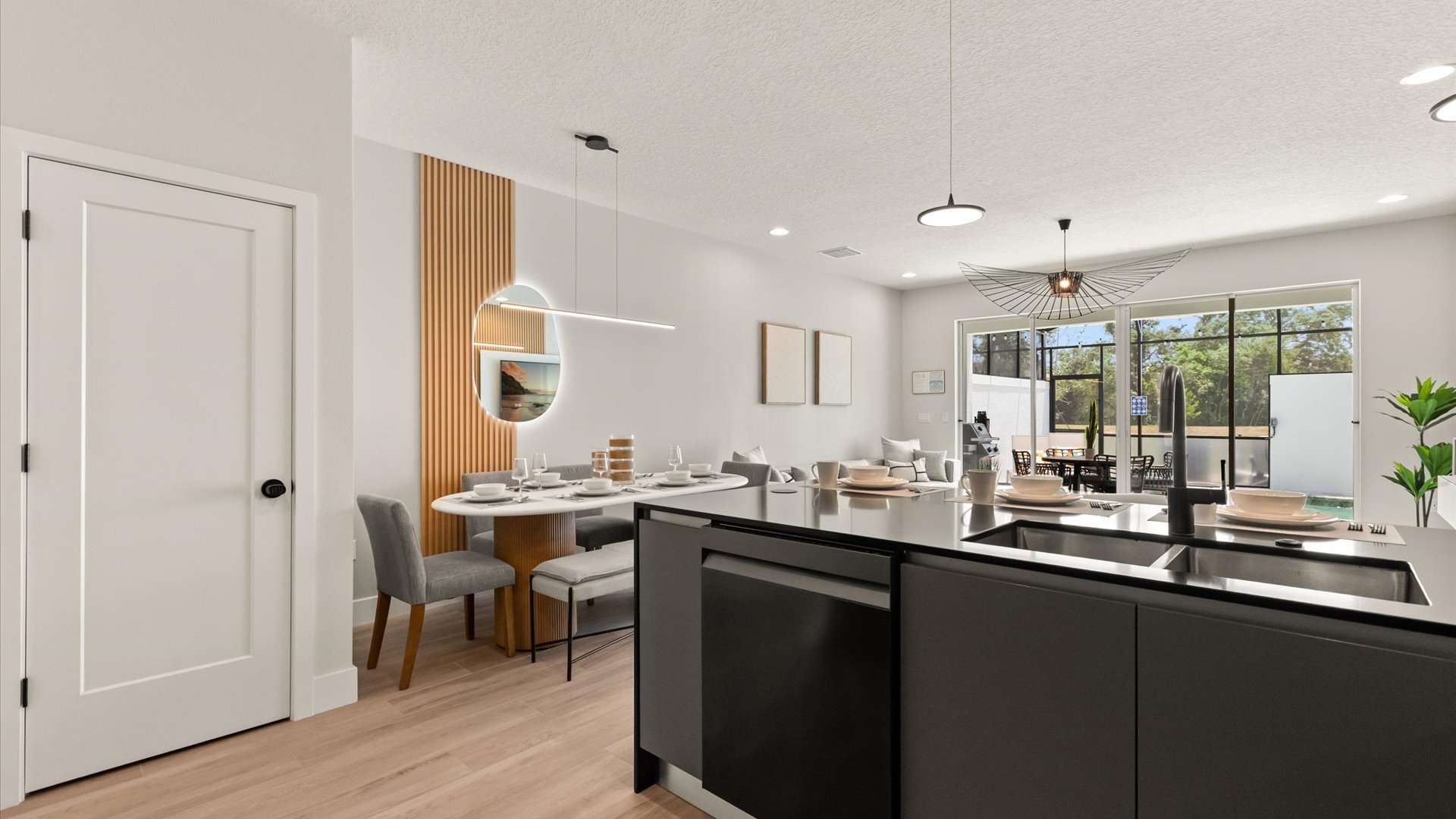 An expansive open-concept space showcasing a dark gray kitchen island in the foreground, a modern dining area with an oval table and bench, and a bright living room visible through to an outdoor area.