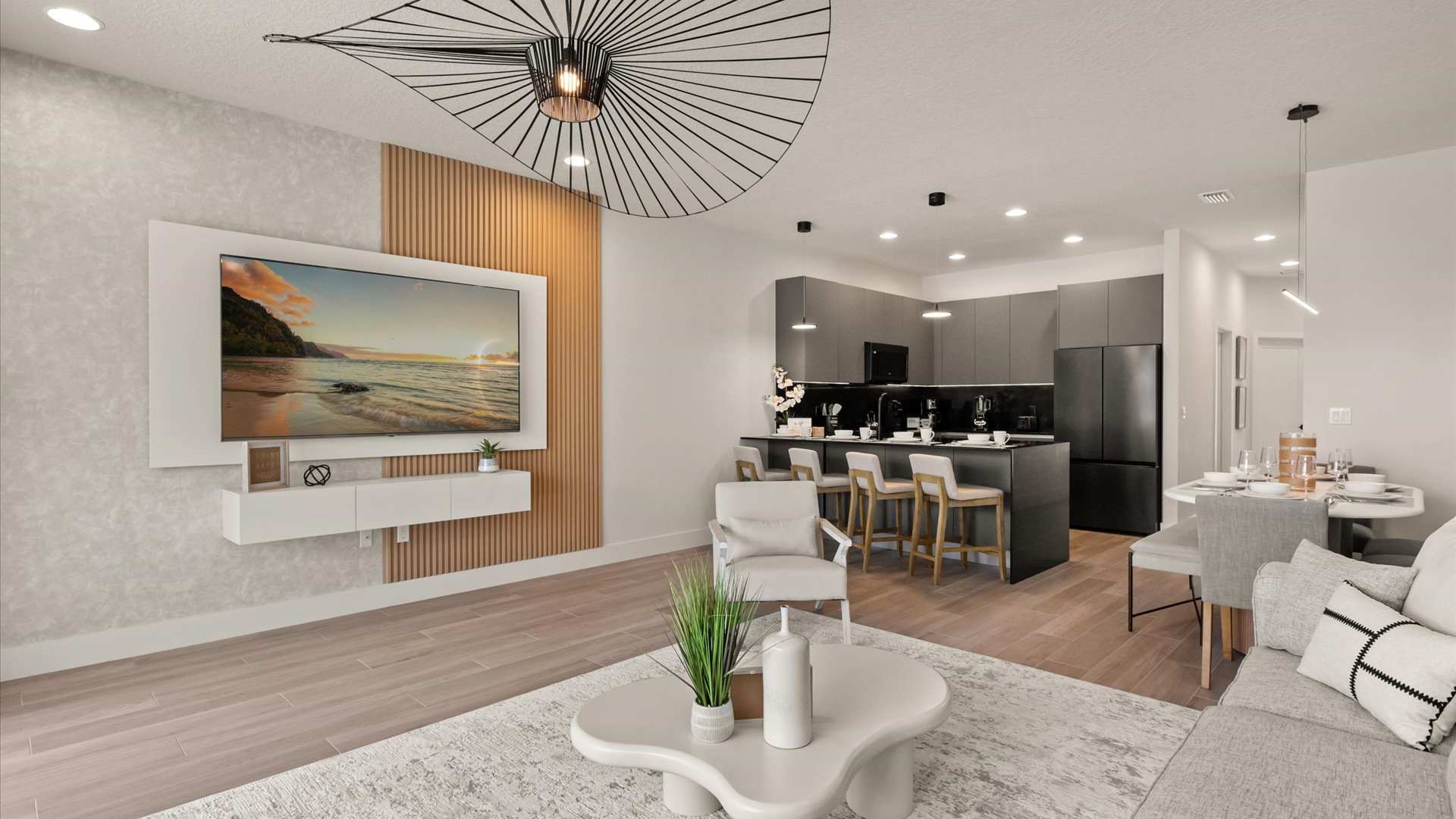 A modern open-concept living room features a large wall-mounted TV above a white floating console, light wood slat accent wall, and a unique black wire chandelier, with a view into the dark gray kitchen and dining area.