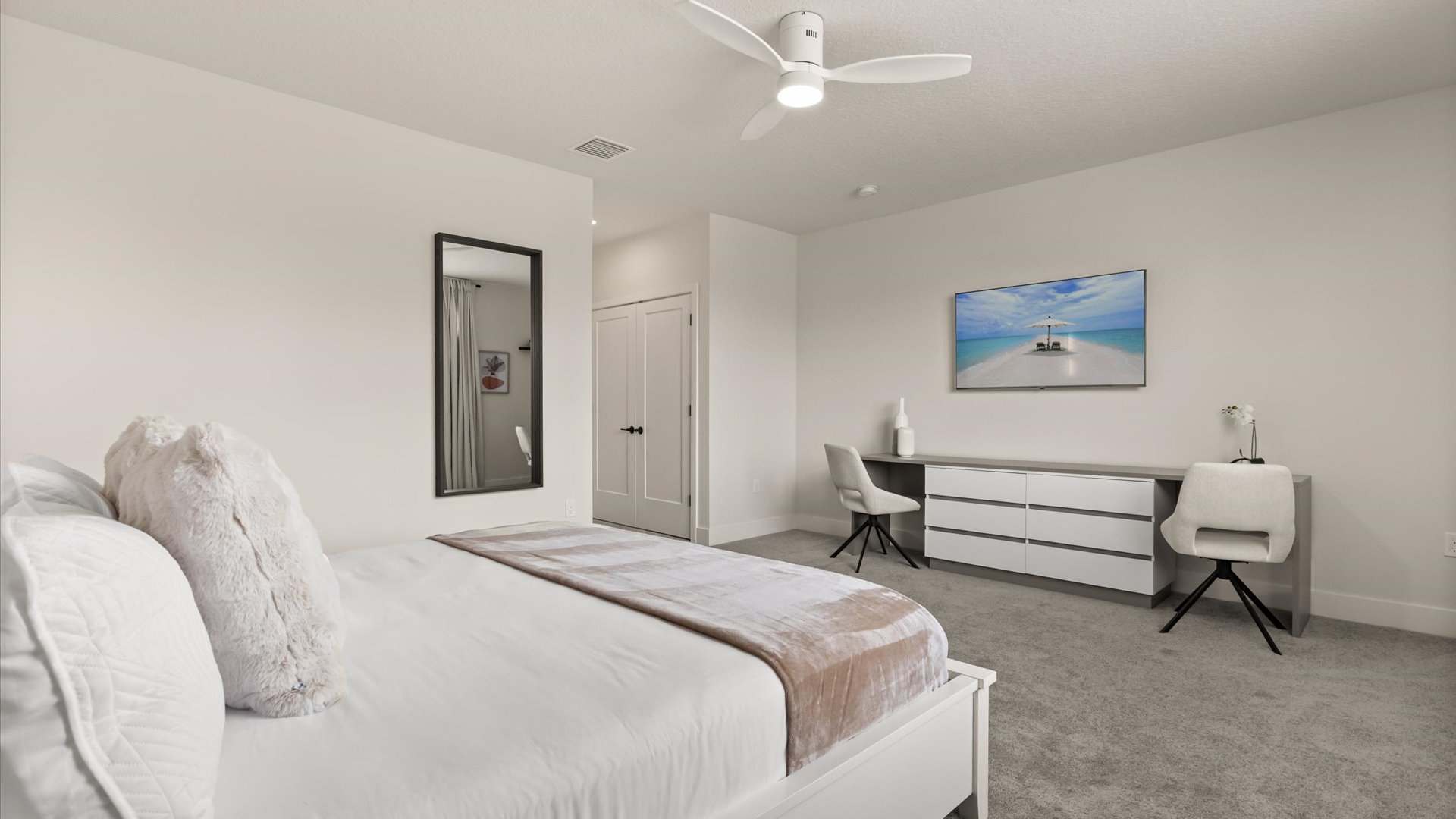 A bright and minimalist bedroom featuring a white bed with plush pillows, a wall-mounted TV above a long gray and white dresser, and two white office chairs, all set on light gray carpet.