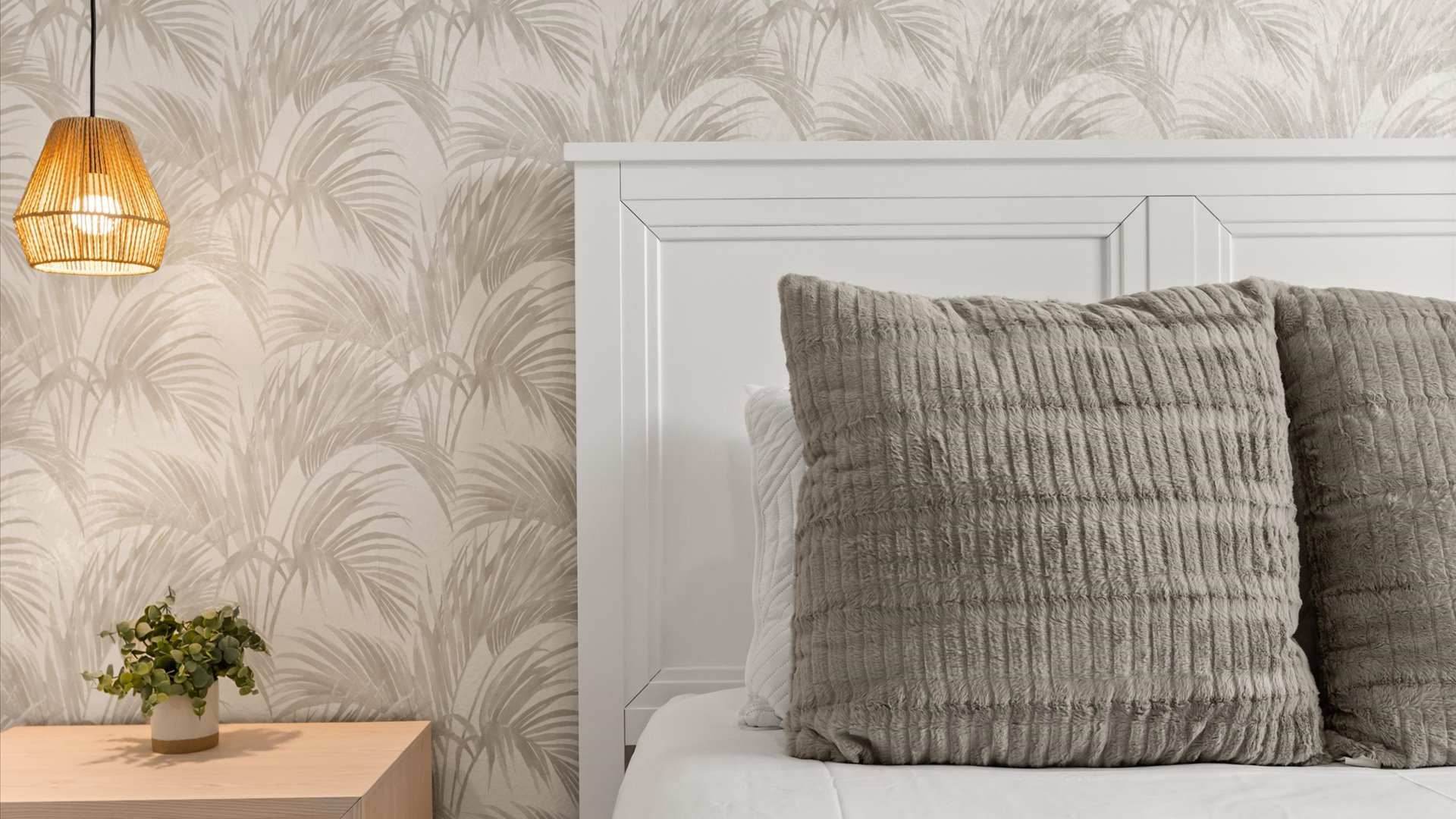Close-up view of a white wooden headboard with a textured gray pillow, a light wood nightstand, and a woven pendant light, set against a subtle palm frond patterned wallpaper.