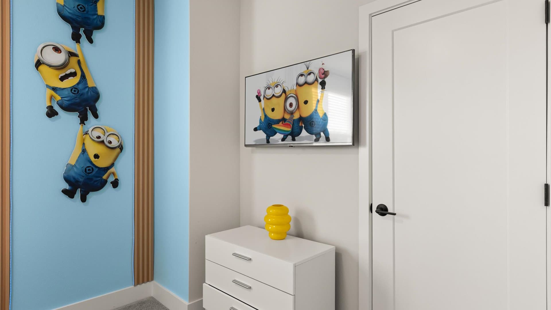 Corner of a kids' bedroom with bright blue walls, Minion wall decor, a white dresser, and a wall-mounted TV displaying Minion characters, next to a white door.