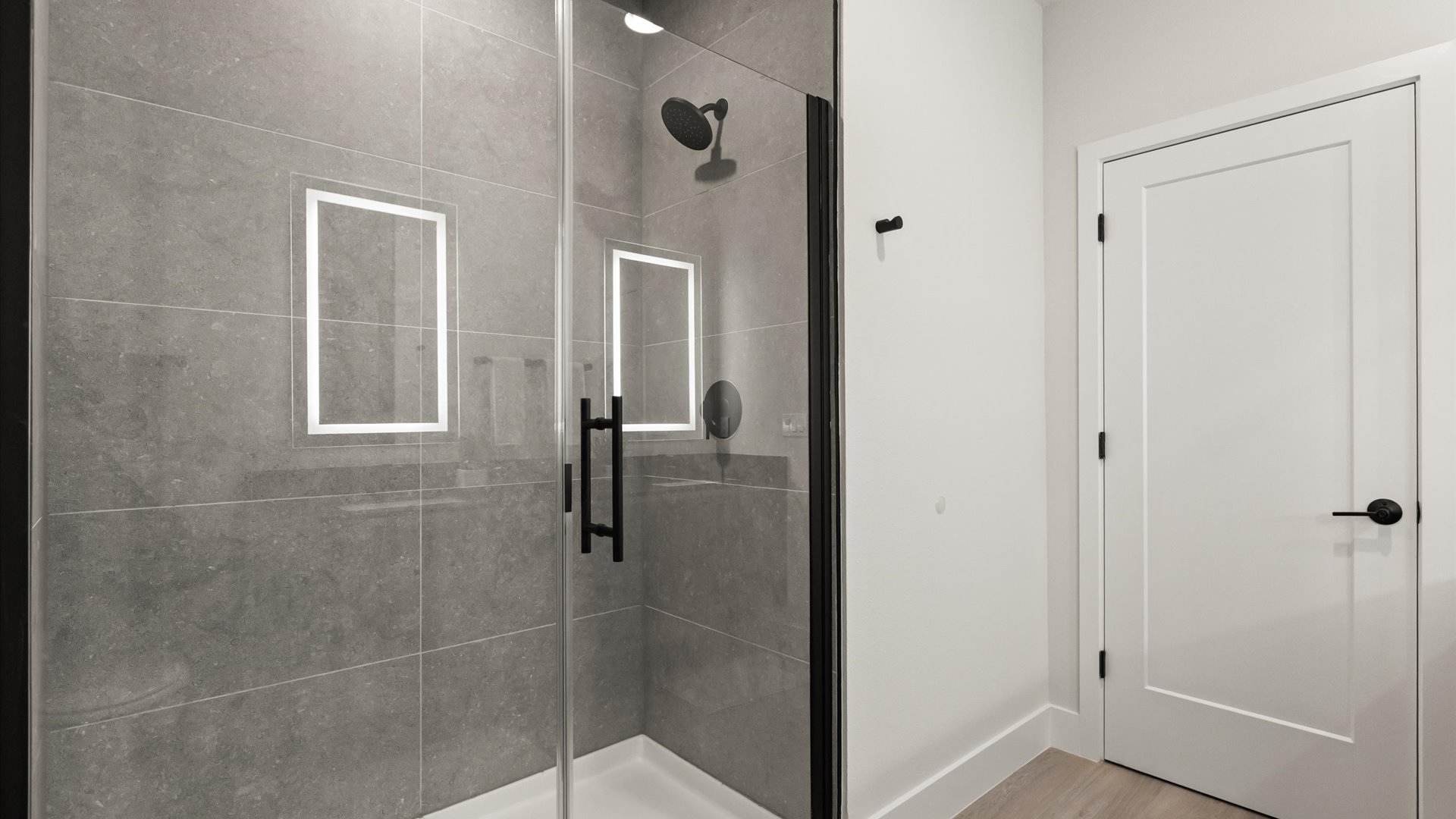 Contemporary walk-in shower with gray subway tile walls, a black-framed glass enclosure, and a black rainfall showerhead, featuring two illuminated rectangular niches.