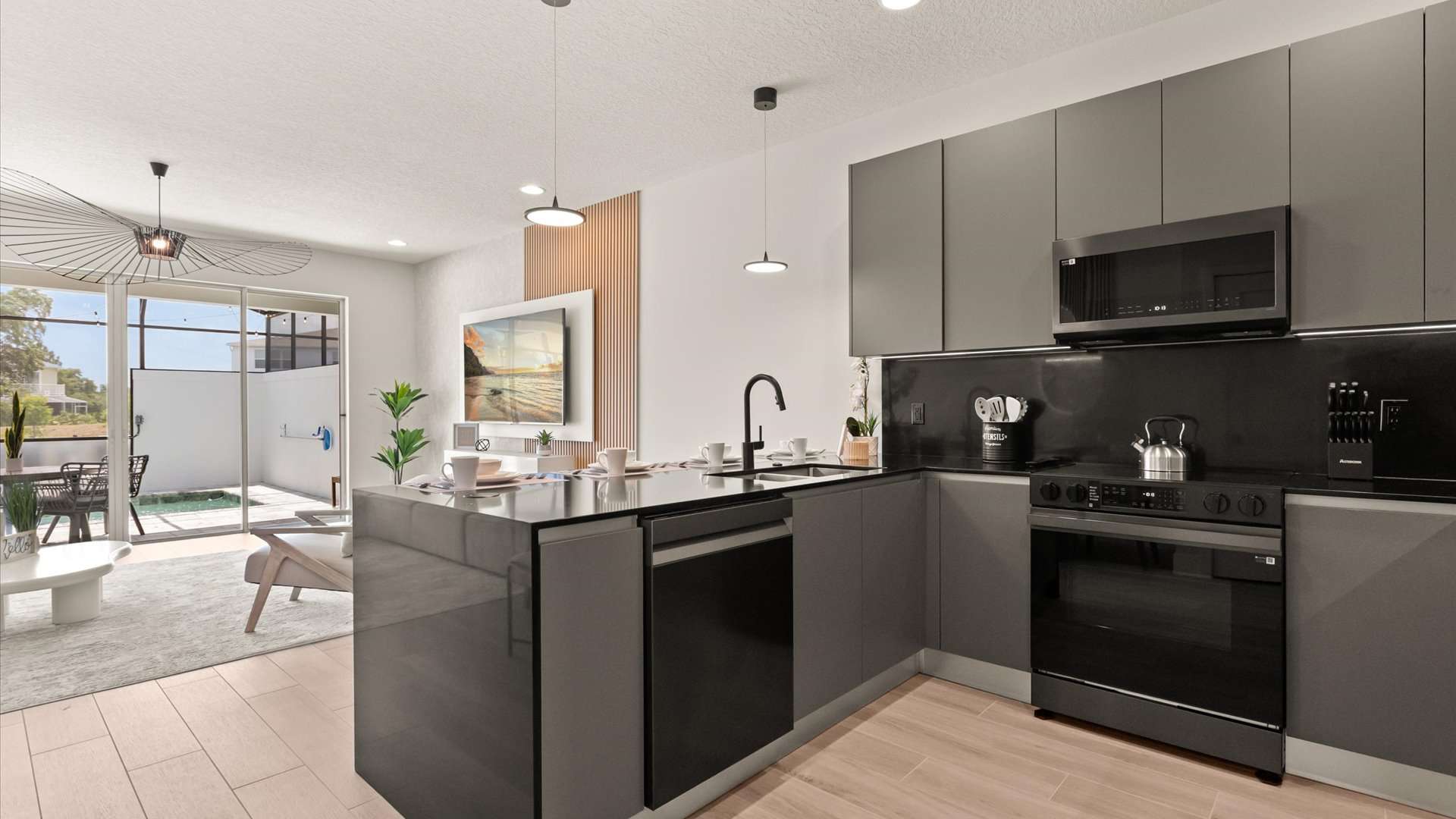 A modern kitchen with dark gray cabinetry and black appliances, featuring a central island with a sink and dishwasher, opening into a living area with a view of an outdoor patio through large sliding glass doors.