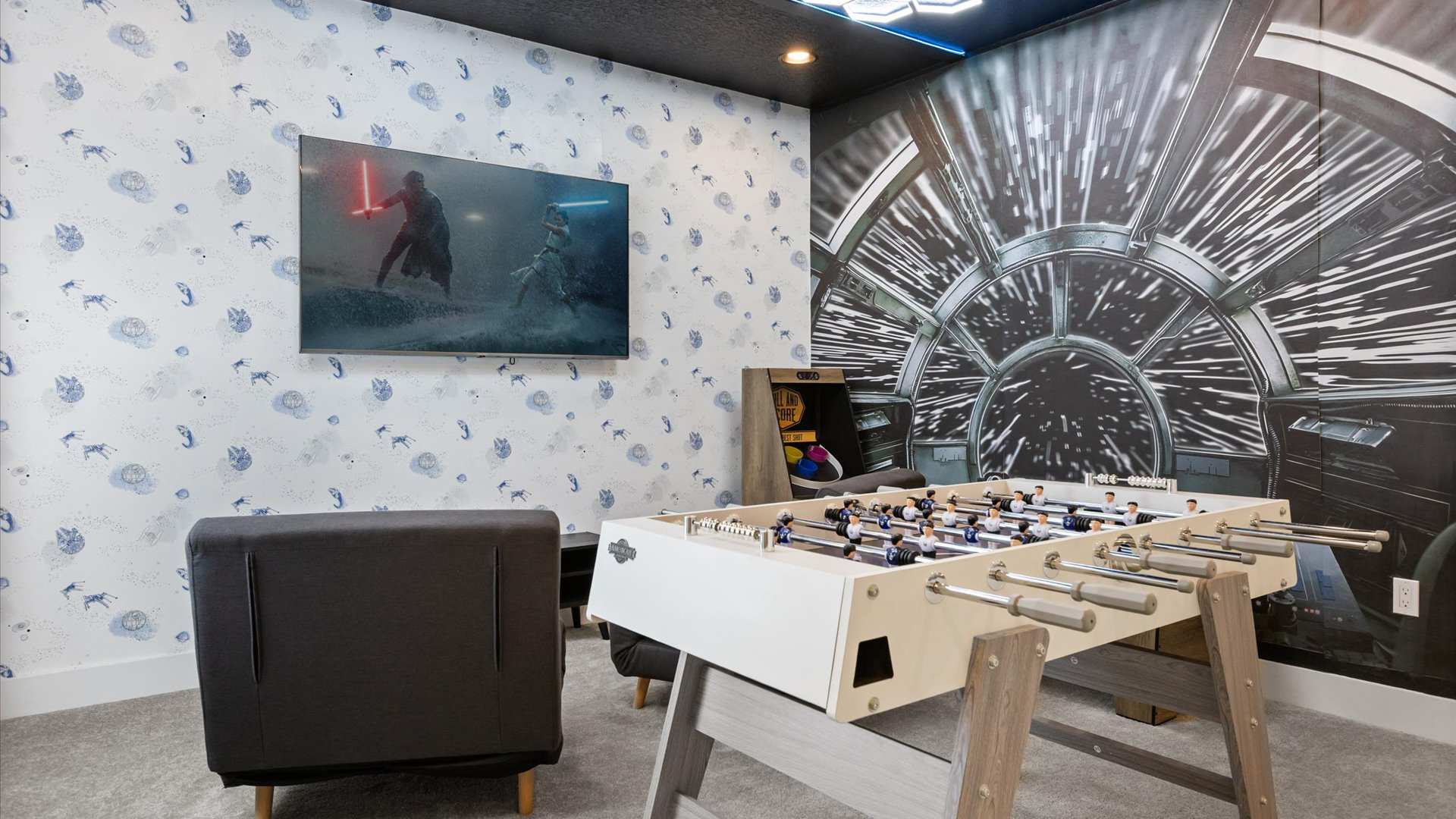 Dynamic game room featuring a large wall mural depicting a spaceship cockpit, a foosball table, and a wall-mounted TV. The room has dark blue walls and a carpeted floor, designed for entertainment.
