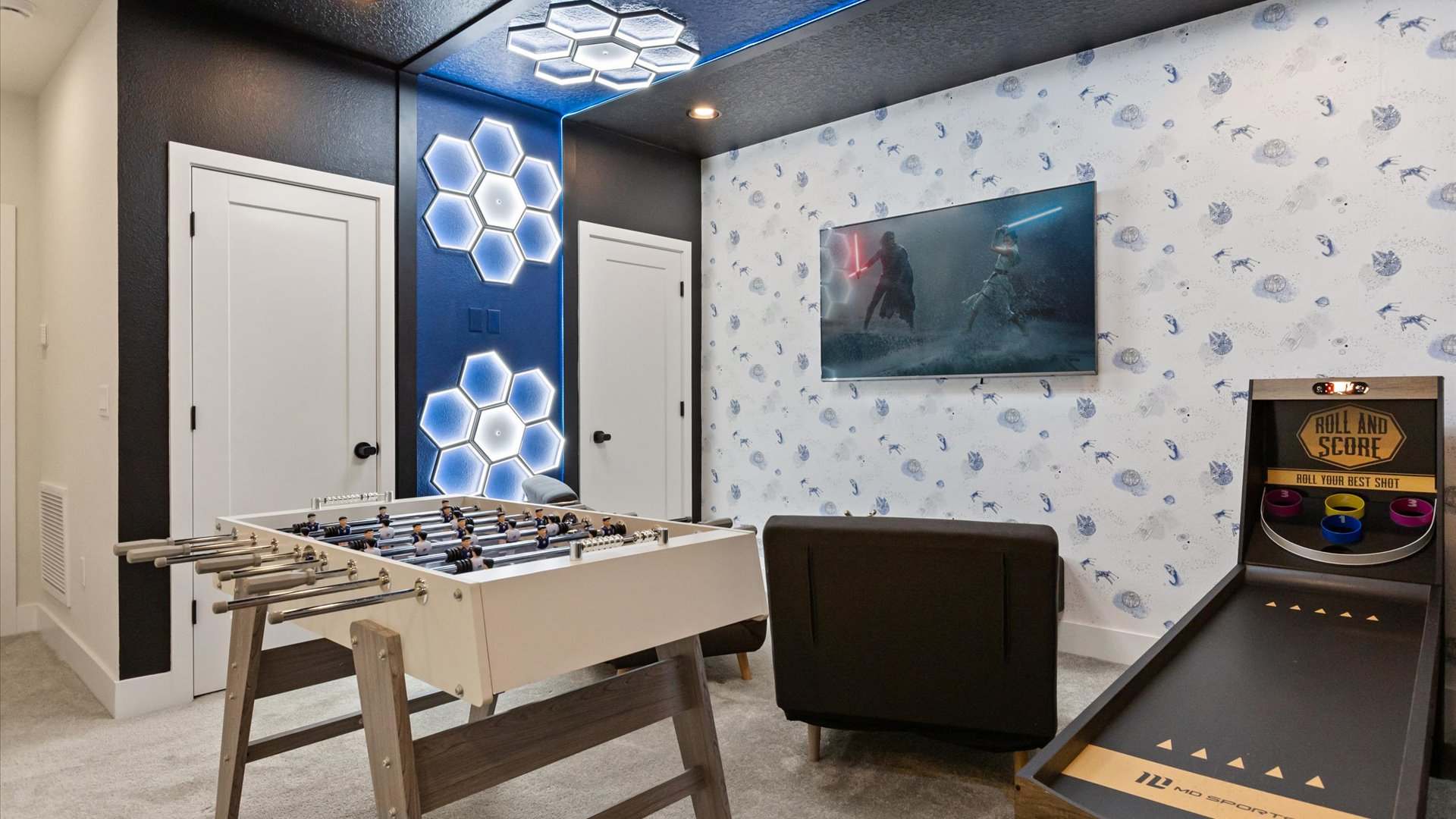 Vibrant game room with a foosball table, a wall-mounted TV displaying a Star Wars scene, and playful blue and white patterned wallpaper. Hexagonal LED lights adorn the dark blue accent wall, creating a dynamic atmosphere.