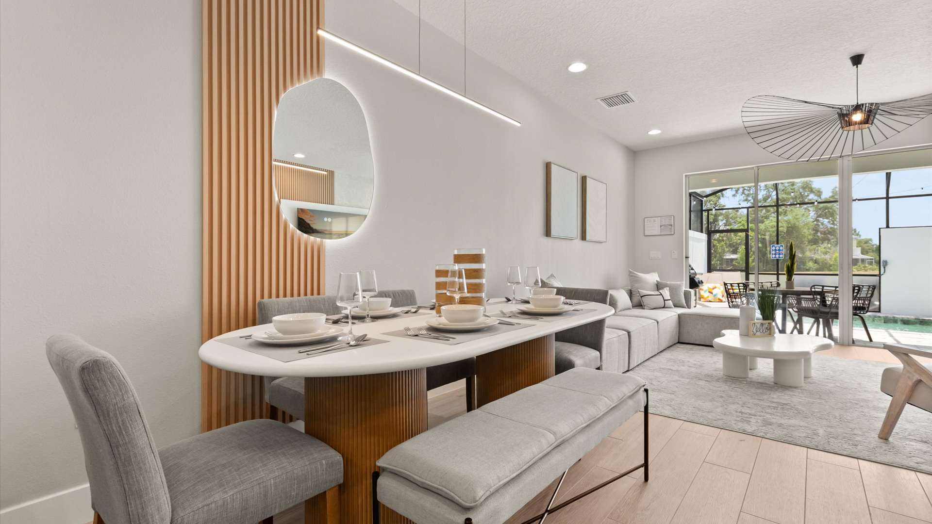 Contemporary dining area featuring an oval white table with a slatted wood base, accompanied by gray upholstered chairs and a matching bench. A long linear pendant light hangs above, and a large oval mirror reflects the space.