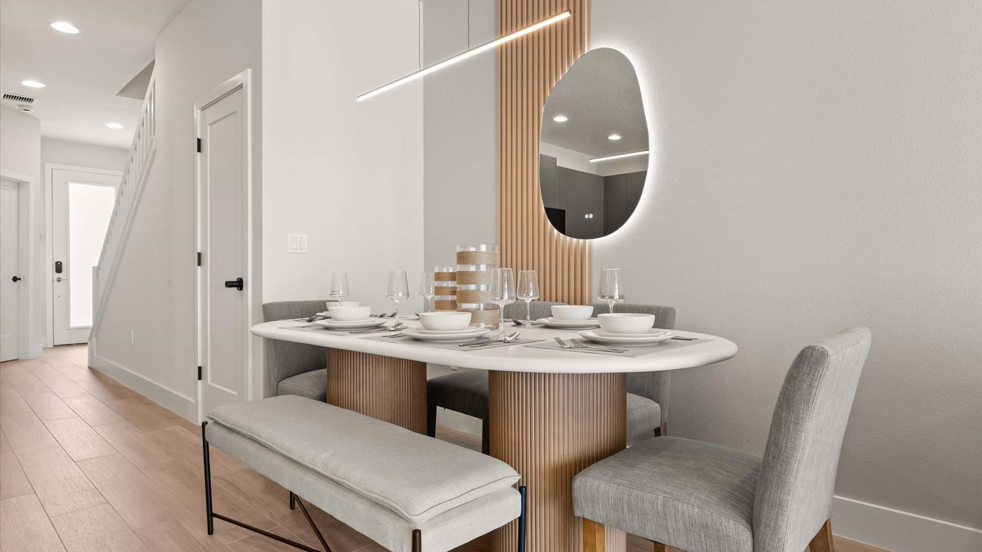 A contemporary dining area with an oval white table supported by fluted wood bases, accompanied by a light gray upholstered bench and matching chairs, set against a wood slat accent wall with an illuminated mirror.