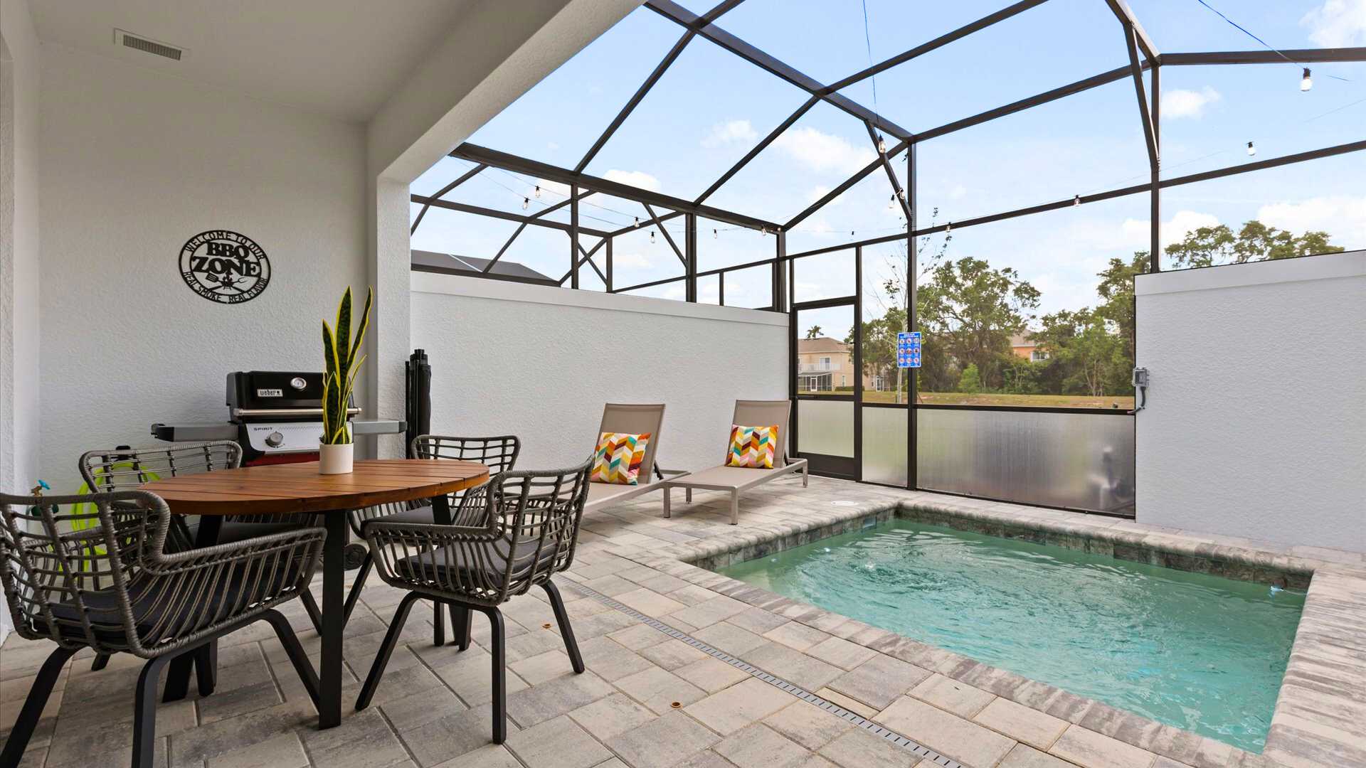 Screened-in outdoor patio featuring a small private plunge pool, two lounge chairs with colorful pillows, and a round dining table with chairs. A BBQ grill is positioned nearby, all set on light-colored pavers.