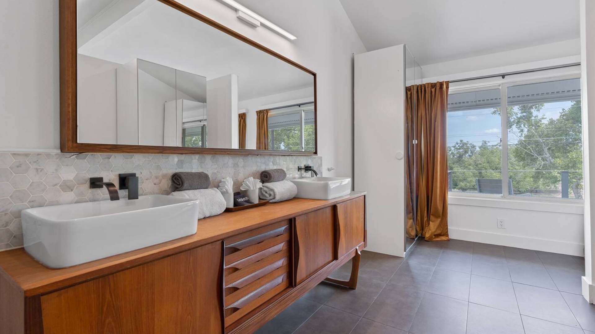Another view of the modern bathroom, highlighting the long wooden double vanity, vessel sinks, and a large window with brown curtains, offering natural light.