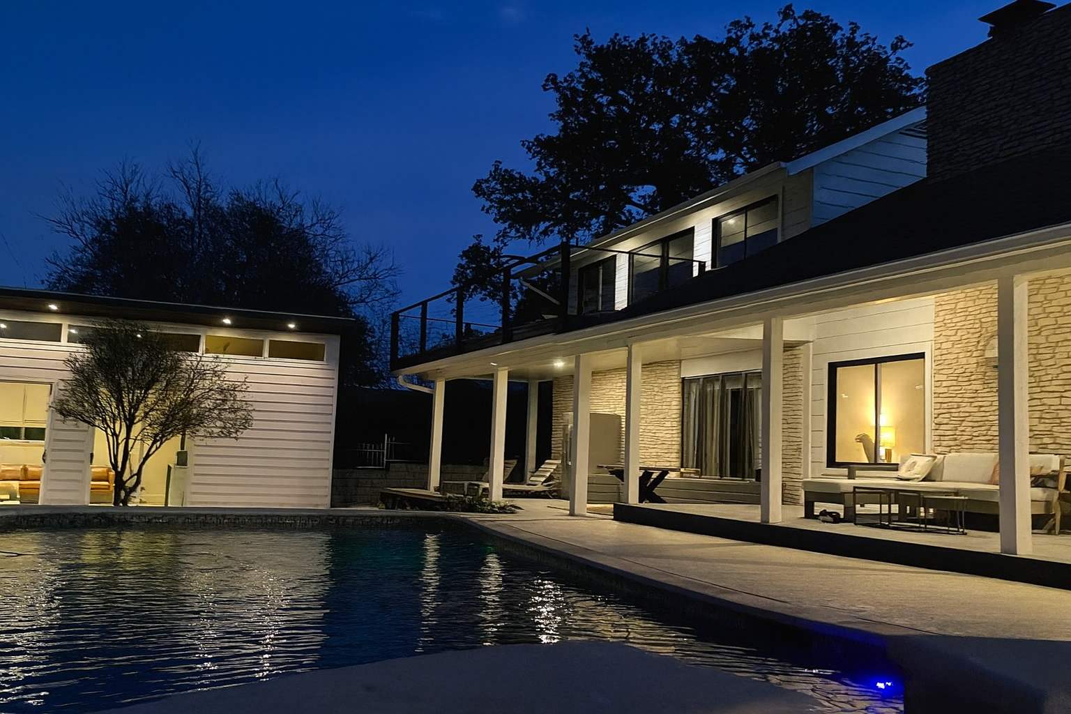 A nighttime view of the house exterior and pool area, illuminated by outdoor lights, showing the sparkling blue pool water and a covered patio with seating, all under a dark blue sky.