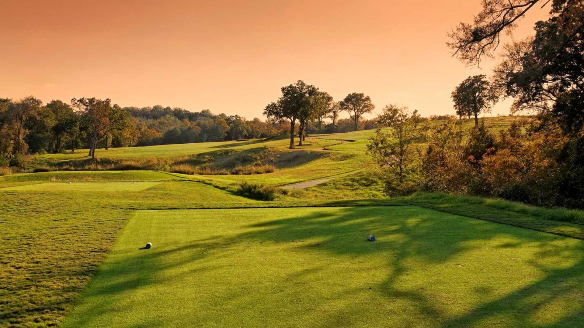 A scenic view of a lush green golf course with rolling hills and scattered trees, bathed in the warm glow of a sunset or sunrise, highlighting the natural landscape.