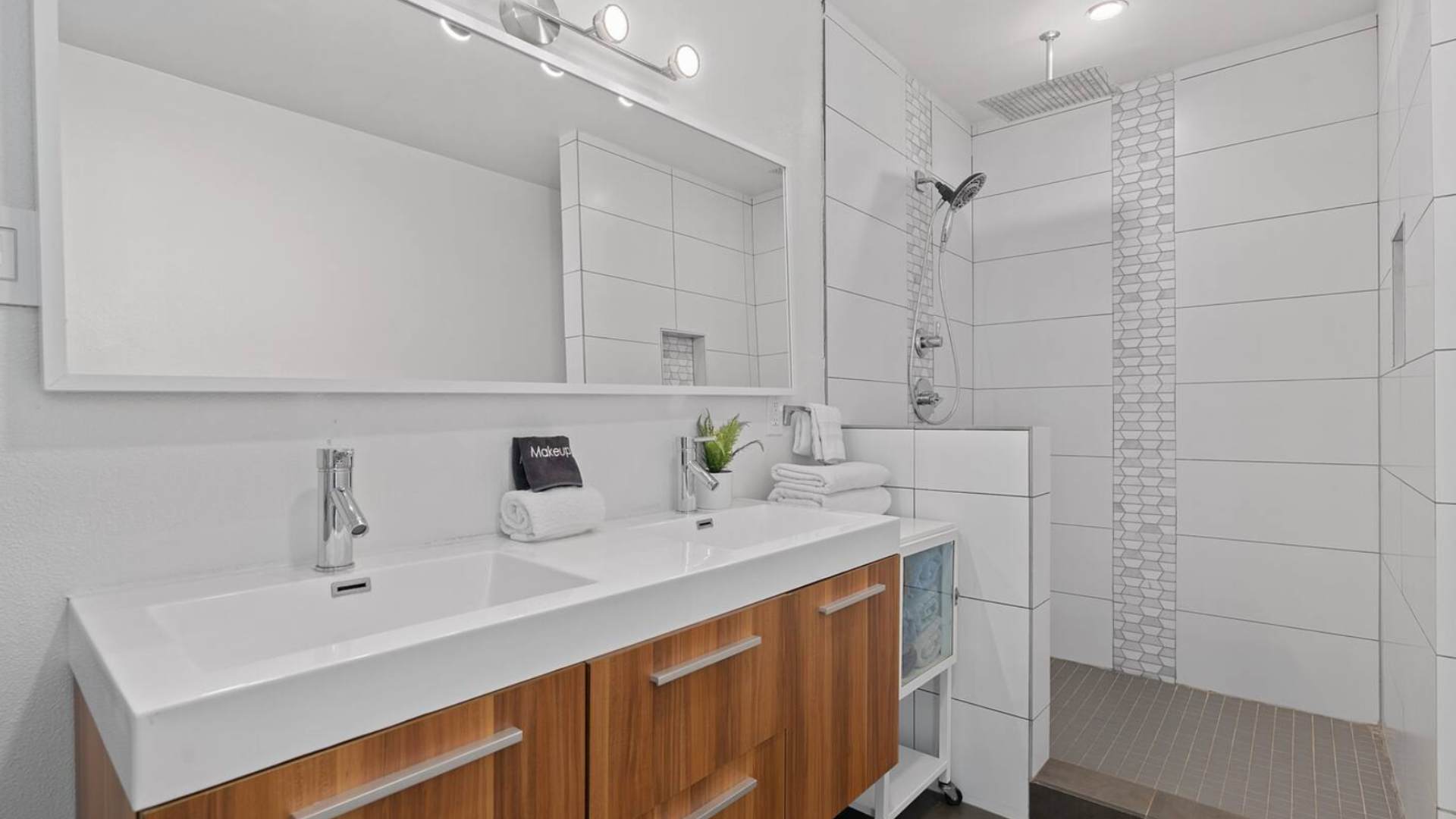 Another perspective of the modern bathroom, showing the double vanity with white sinks, a large mirror, and the walk-in shower with white and mosaic tiling.