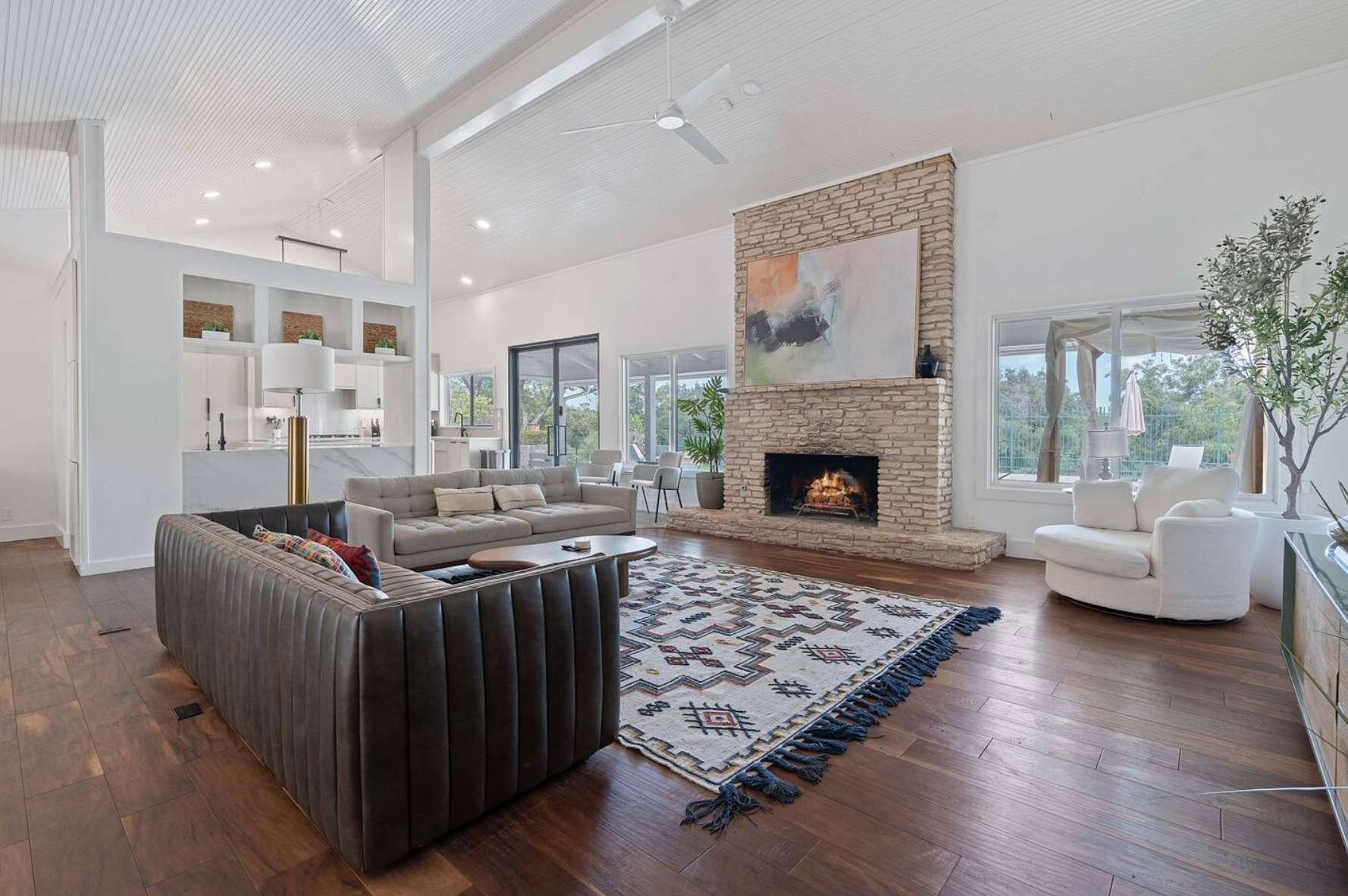 Spacious living room featuring a prominent stone fireplace with a large abstract painting above, a light grey sofa, and a dark leather sectional, all on a patterned area rug with dark hardwood floors.