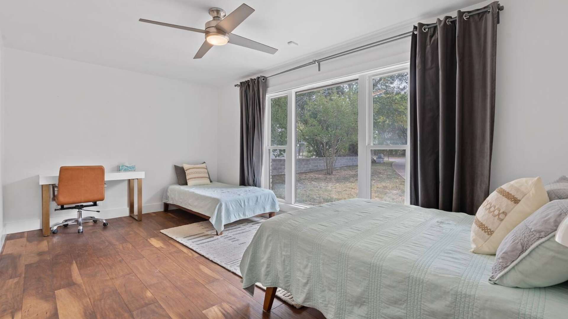 A bright bedroom with two beds, one larger and one twin, featuring a modern desk with an orange office chair, large windows with dark curtains, and a ceiling fan.