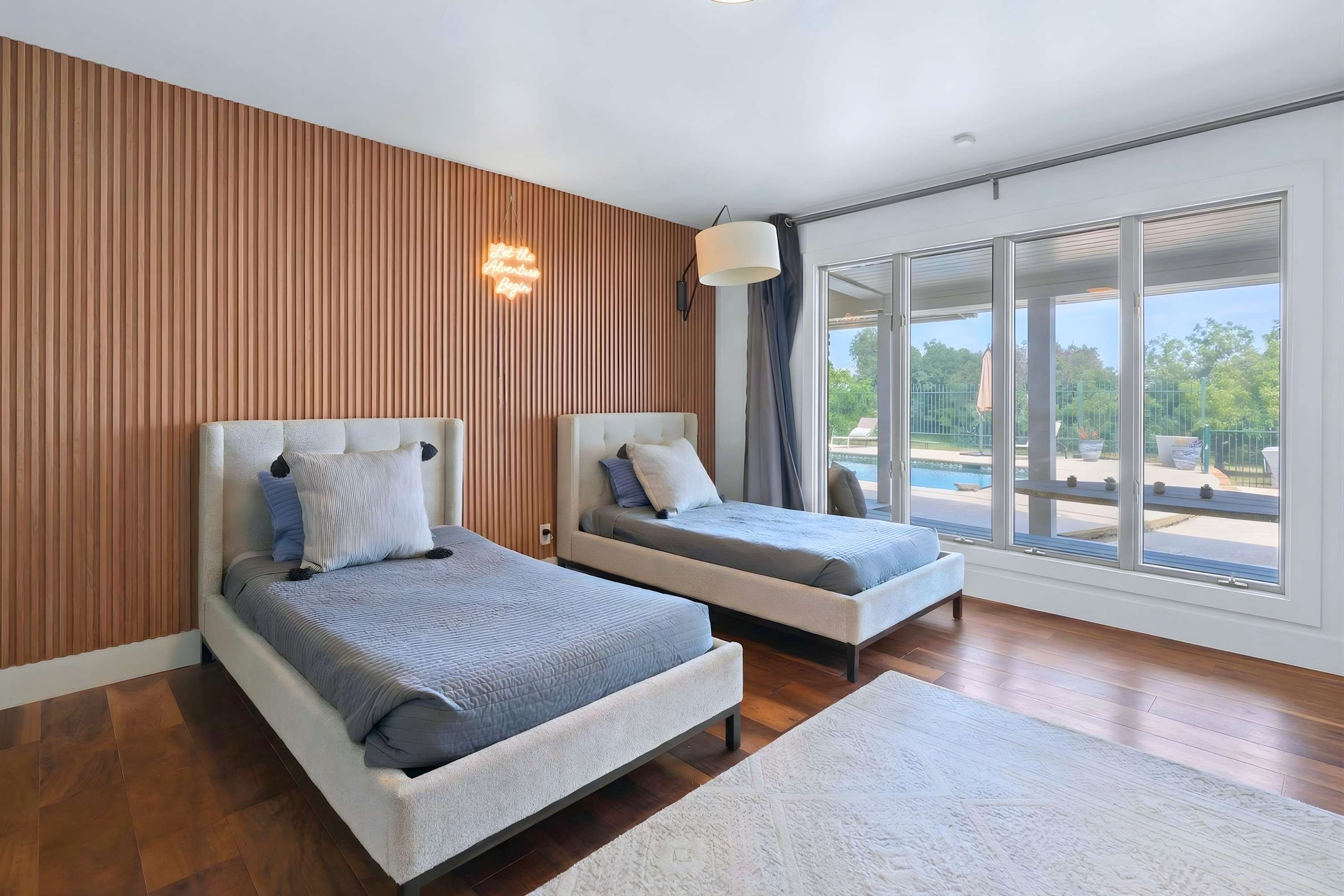 A modern bedroom featuring two twin beds with upholstered headboards, a striking slatted wooden accent wall with a neon sign, and large windows offering a view of an outdoor pool.