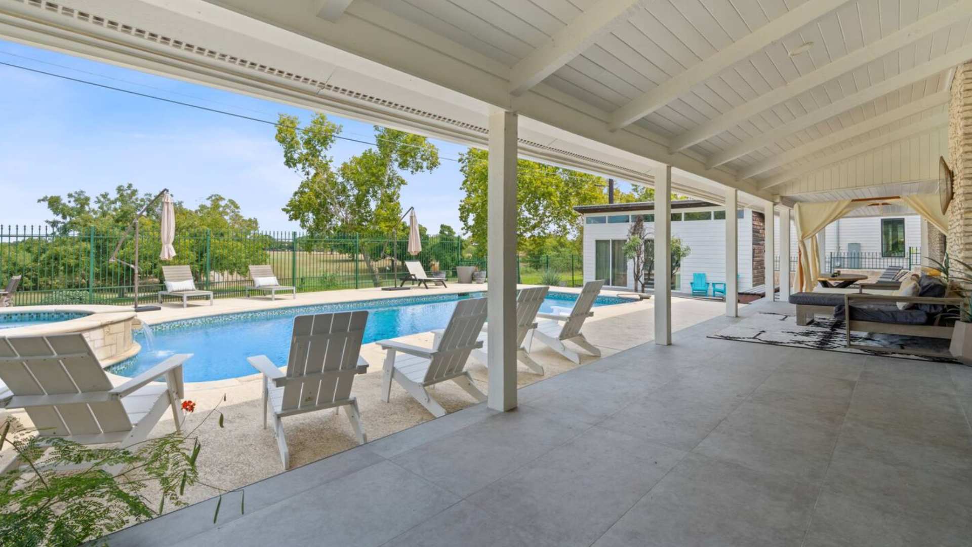 A covered patio area with white columns overlooks a sparkling blue swimming pool and hot tub, surrounded by white Adirondack chairs and lush greenery, with a golf course visible in the distance.
