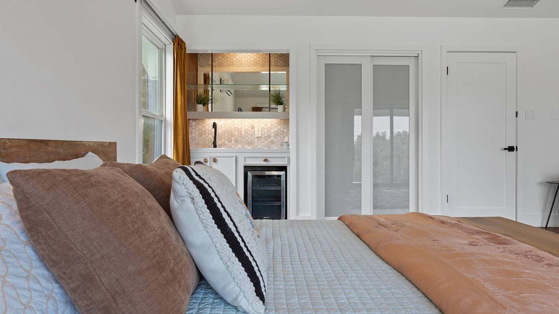 Close-up of the bedroom's wet bar area, featuring a small sink, open glass shelving, a mini-fridge, and a stylish hexagonal tile backsplash.