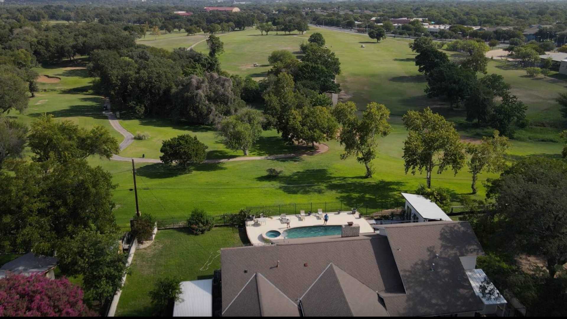 An aerial view showcases a large house with a brown roof and a blue swimming pool, situated beside a sprawling green golf course with numerous trees under a clear sky.