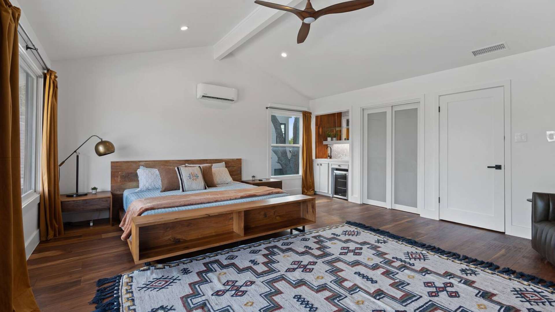 Spacious master bedroom with a wooden platform bed, a large patterned rug, a ceiling fan, and a built-in wet bar with a mini-fridge and open shelving.