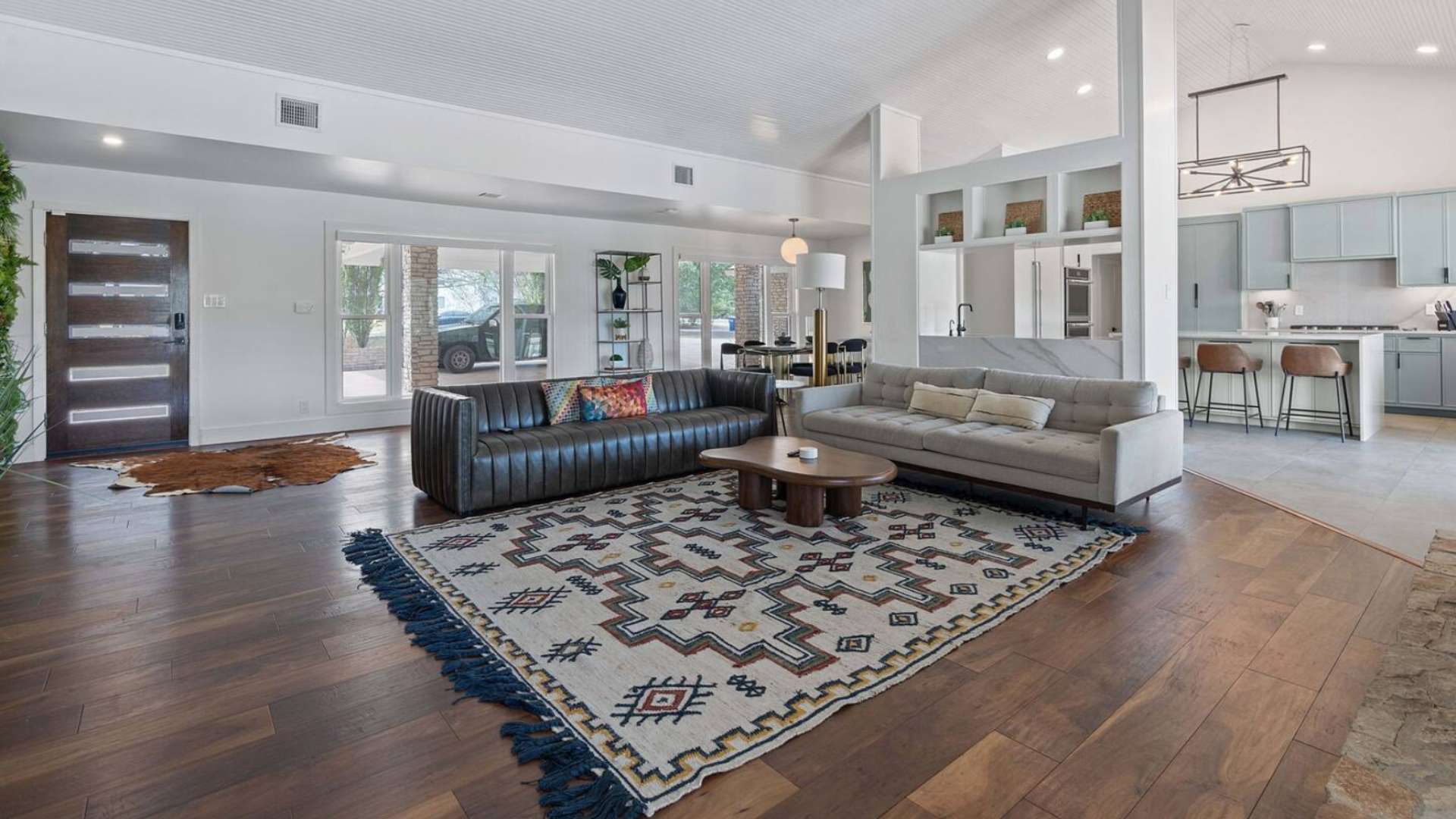 An open-concept living area with dark wood floors and a large patterned area rug, featuring a dark leather sofa and a light fabric sofa, leading into a modern kitchen with a large island.
