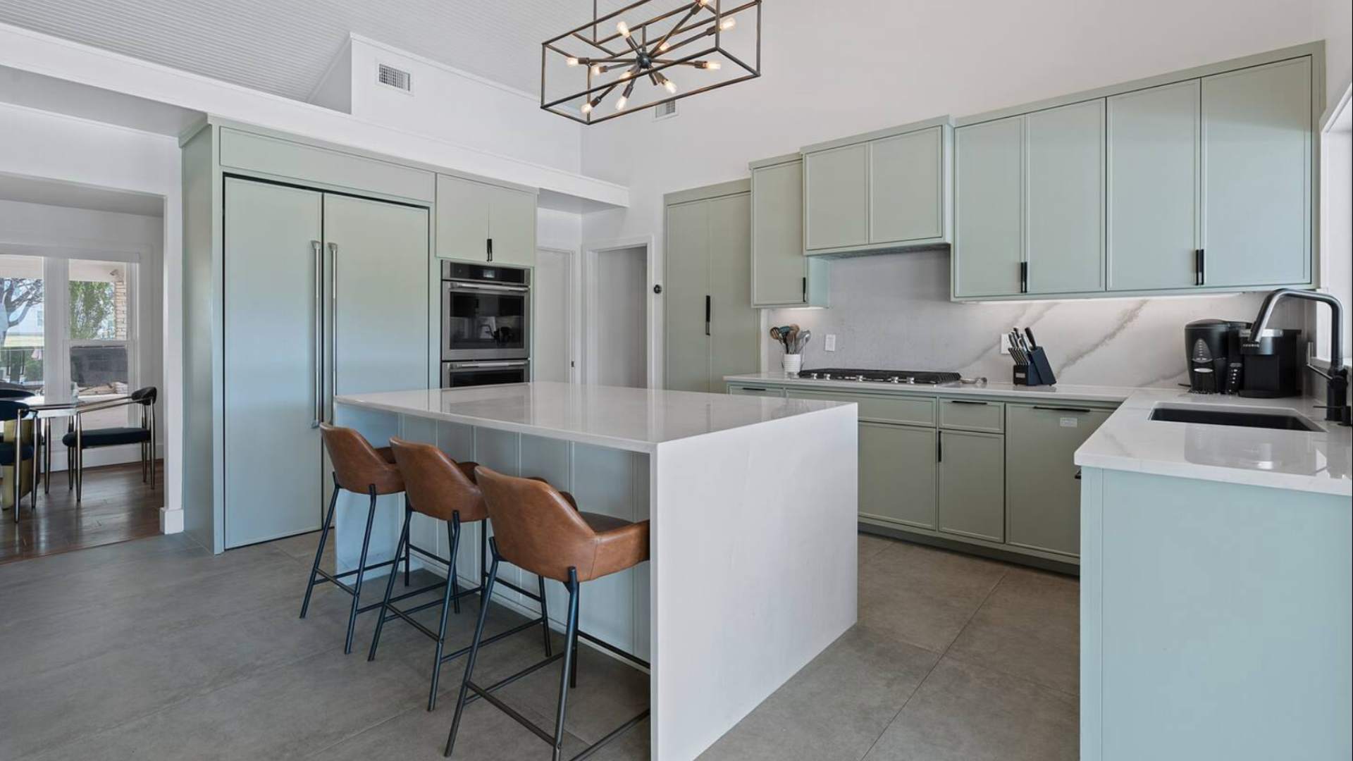 Spacious kitchen with light green shaker-style cabinetry, integrated refrigerator, a large white island with three leather bar stools, and a modern linear chandelier overhead.