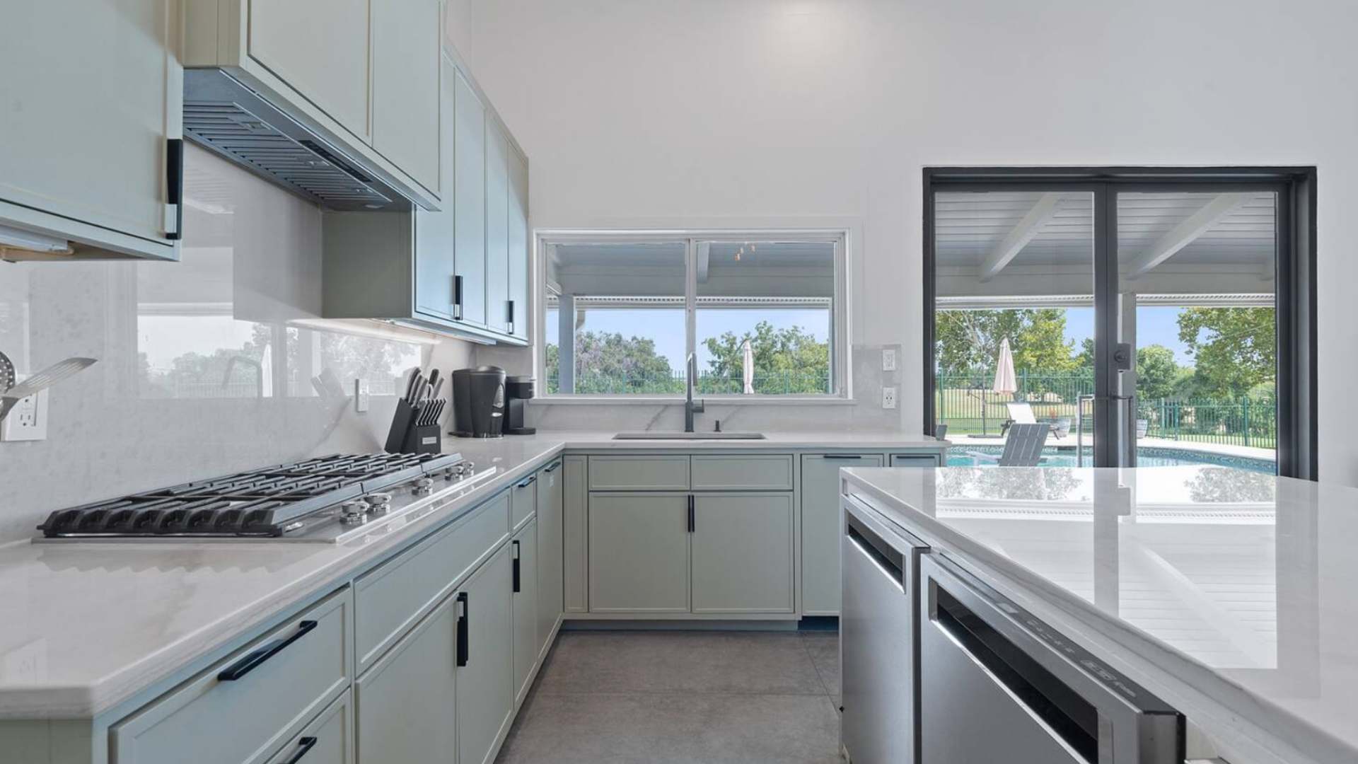 Kitchen counter with a built-in gas range and overhead ventilation, featuring light green cabinetry, white quartz countertops, and a large window overlooking the lush outdoor landscape.
