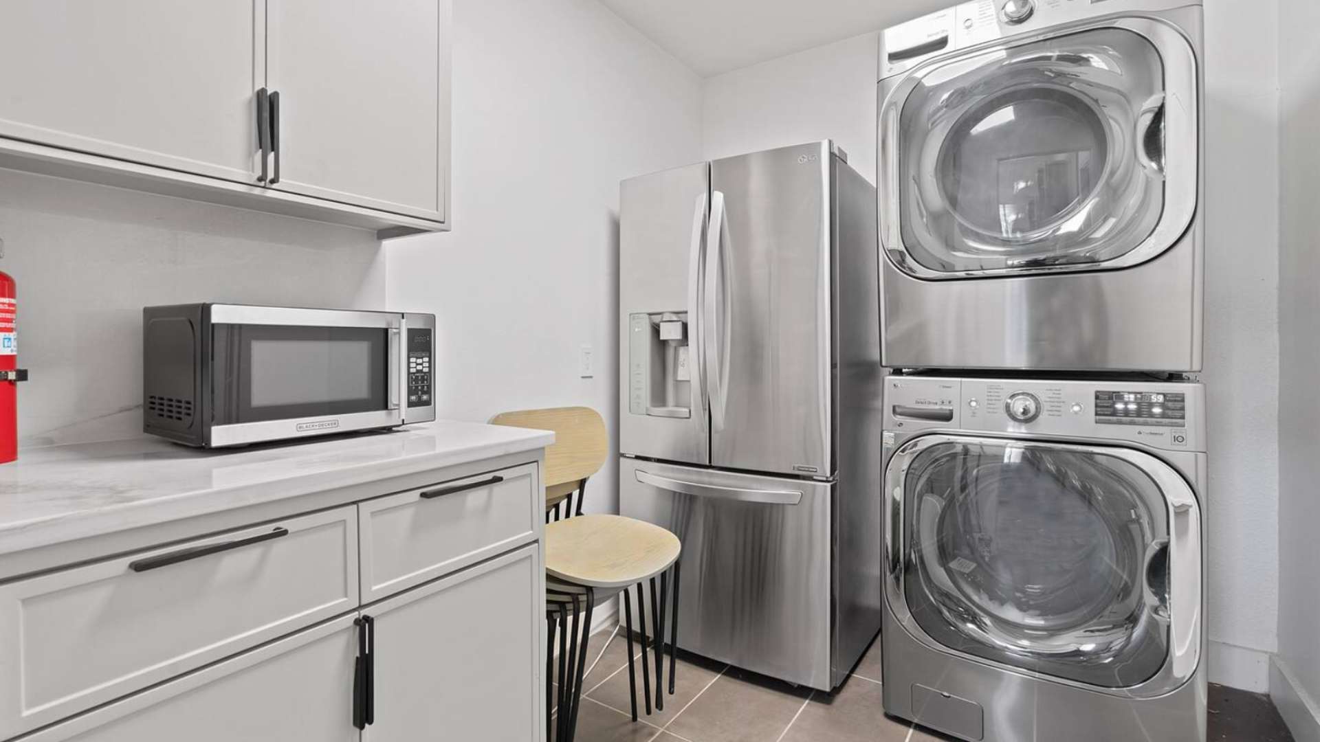 A functional laundry area equipped with a stacked washer and dryer, a full-size stainless steel refrigerator, a microwave, and light gray cabinets with a white countertop.