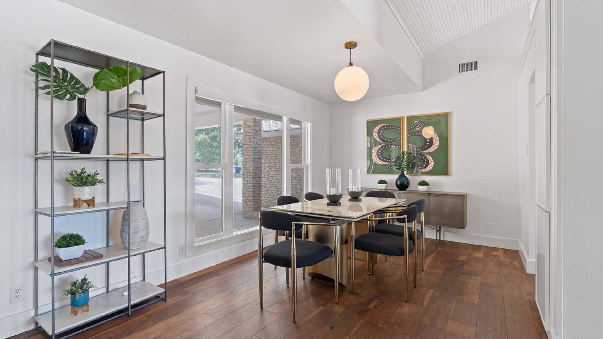 Modern dining area featuring a rectangular table with a gold base and black chairs, illuminated by a spherical pendant light, alongside a tall metal shelving unit adorned with plants and decor.