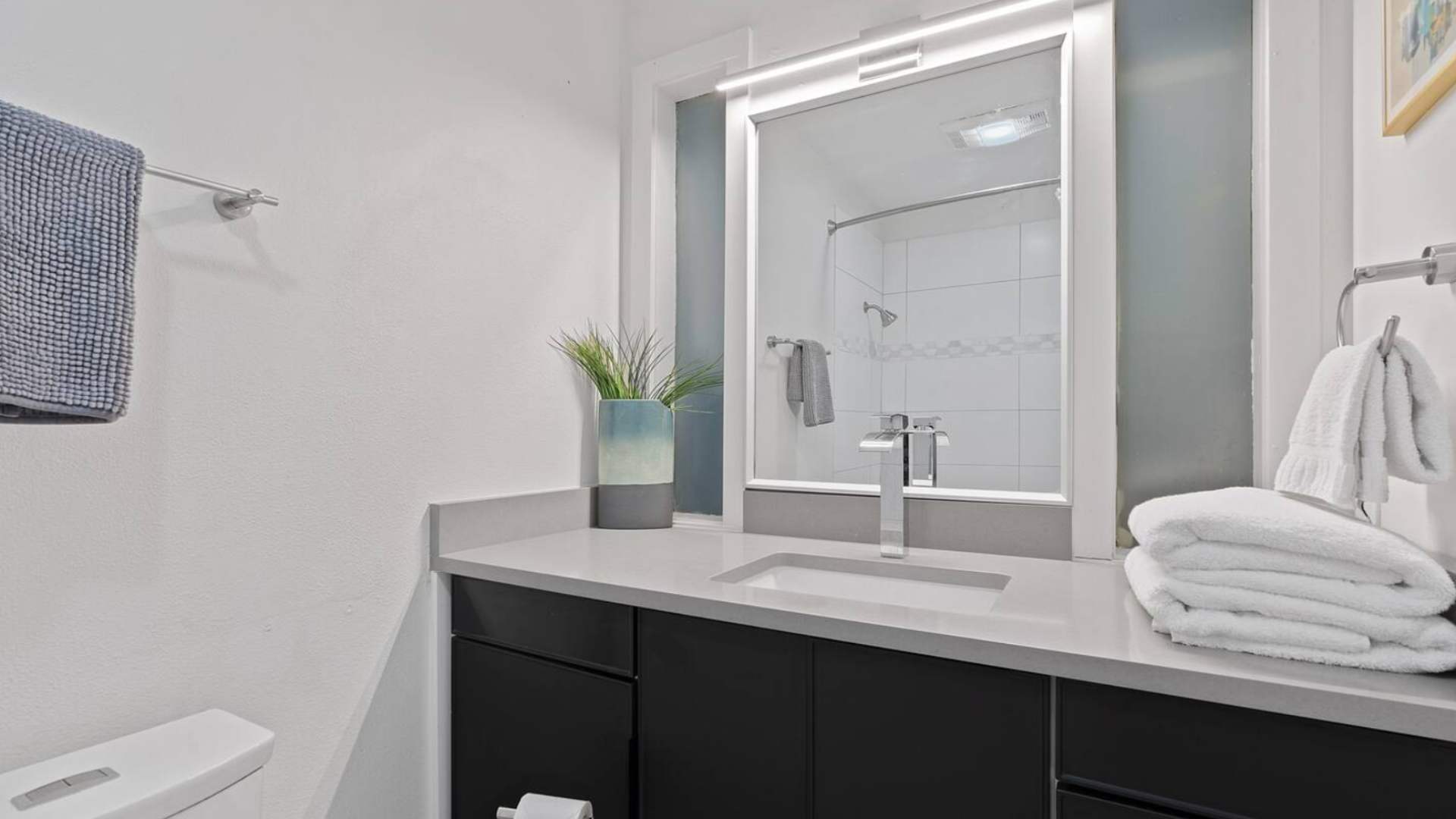 A compact modern bathroom featuring a single vanity with dark cabinetry, a light gray countertop, an illuminated mirror, and a shower with white tiled walls visible in the reflection.