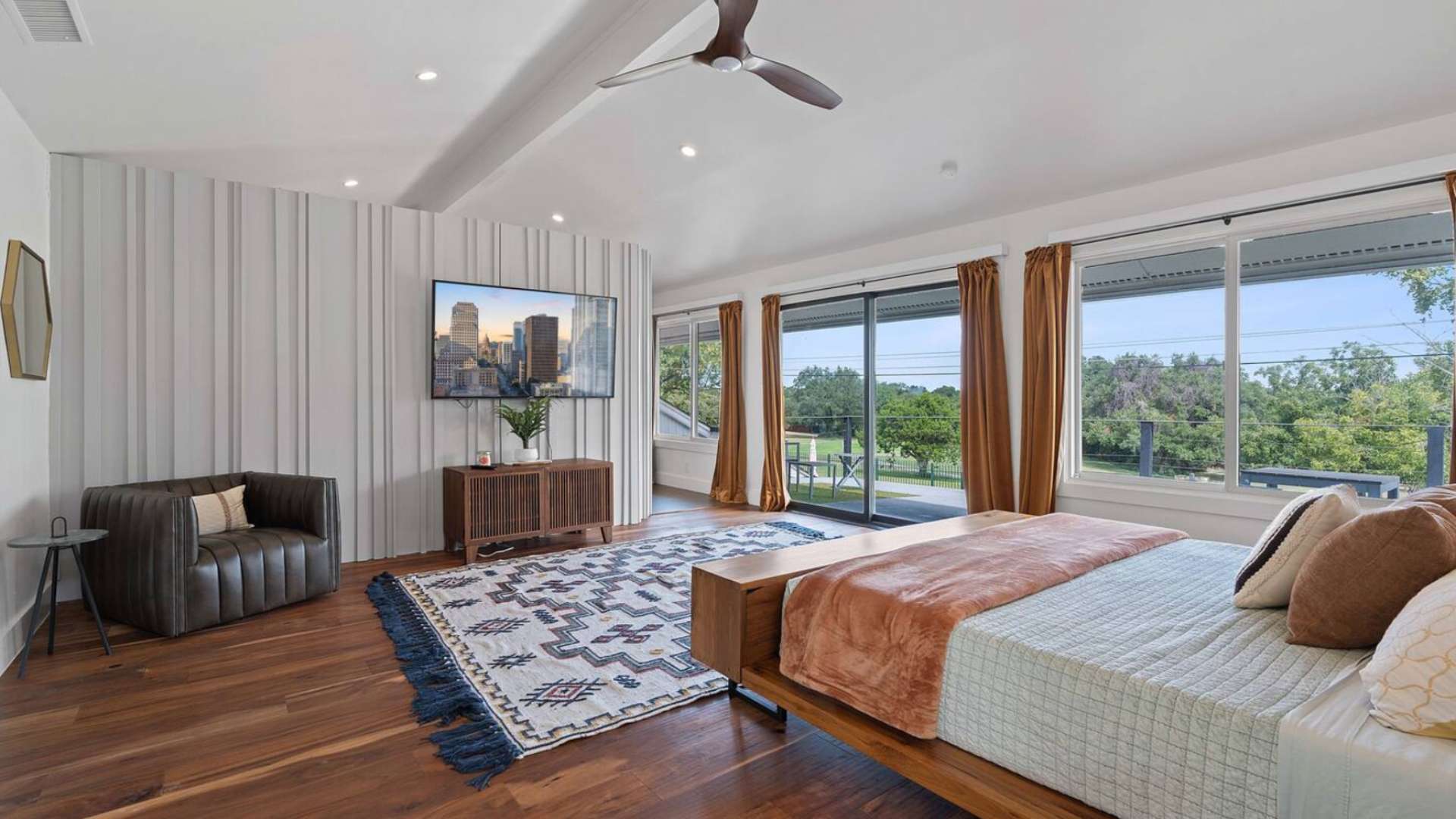 Wide view of the master bedroom featuring a wooden bed, a large patterned rug, a wall-mounted TV on a textured accent wall, and expansive windows overlooking greenery.