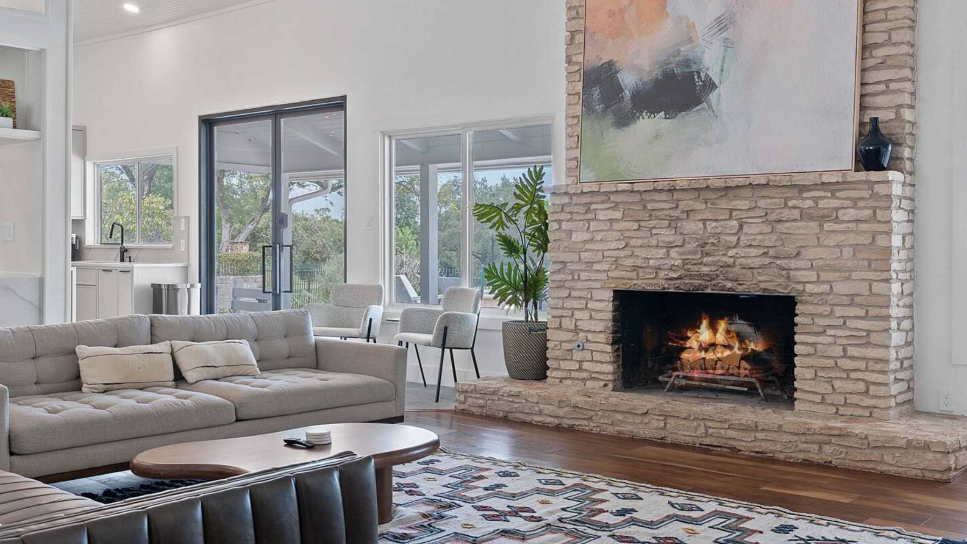Cozy living room corner showcasing a rustic stone fireplace with a roaring fire, a comfortable grey sofa with throw pillows, and large windows offering a view of the outdoor patio and greenery.