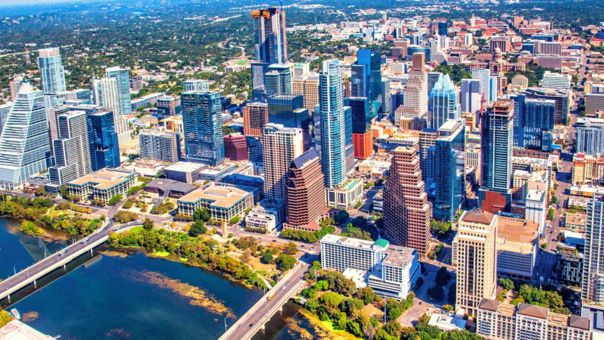 An expansive aerial view of a bustling city skyline with numerous high-rise buildings, situated alongside a wide river with bridges, under a clear blue sky.