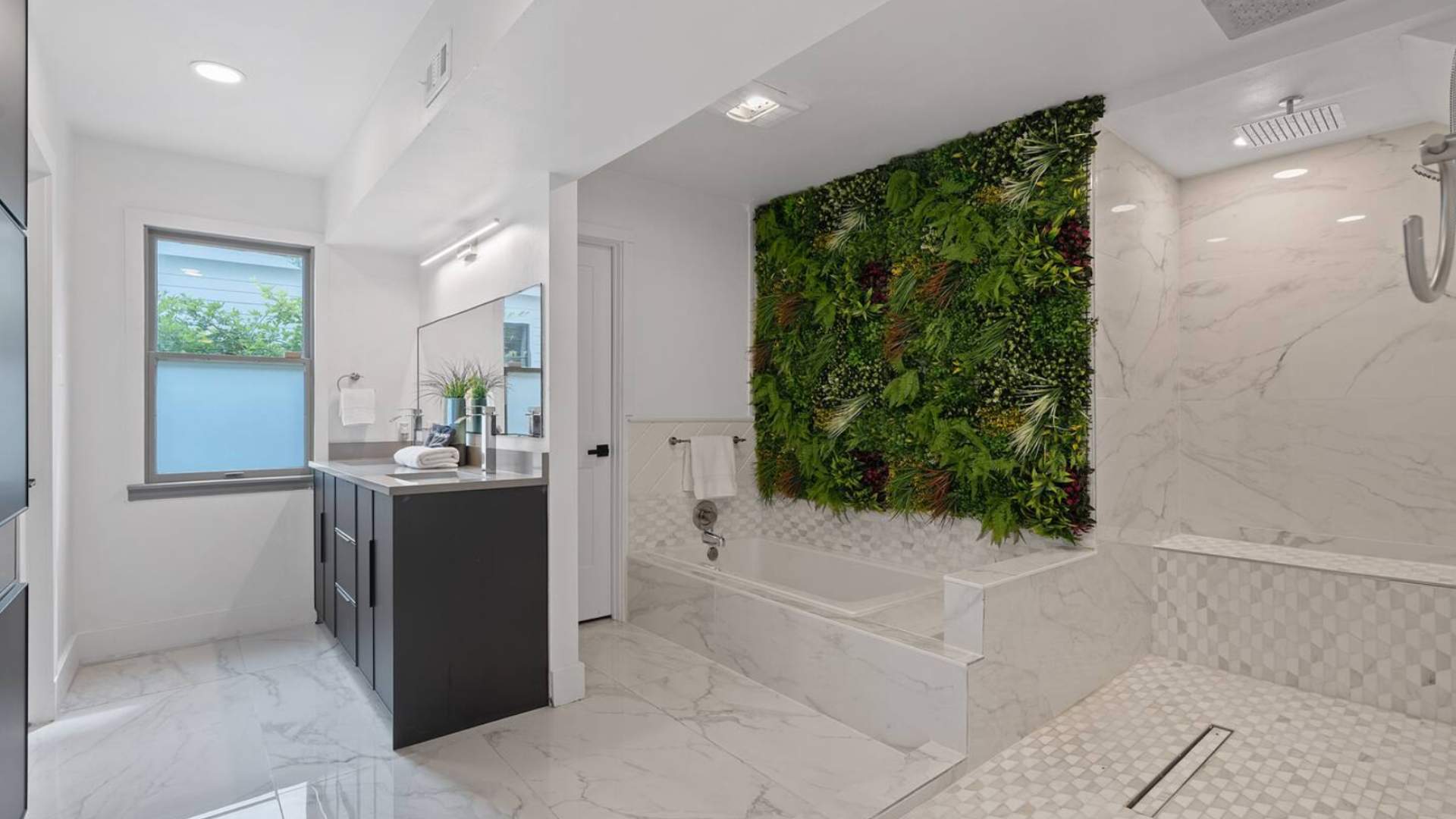 A spacious and modern bathroom showcasing a deep soaking tub with a striking green living wall, a large walk-in shower with marble tiling, and a dark wood vanity with a sleek countertop and illuminated mirror.