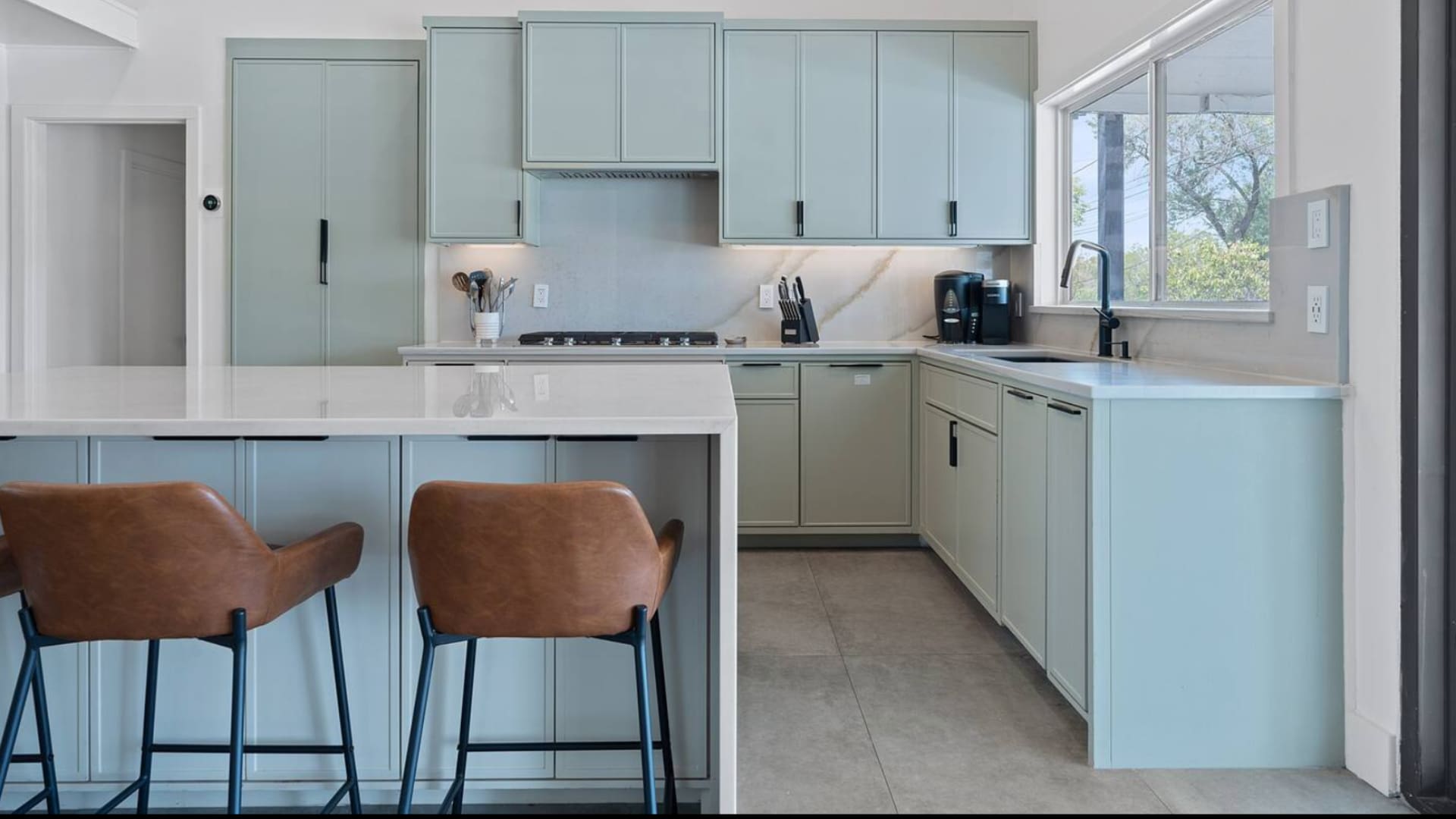 Modern kitchen featuring light sage green cabinetry, white quartz countertops, a marble-patterned backsplash, and a large island with two brown leather bar stools.