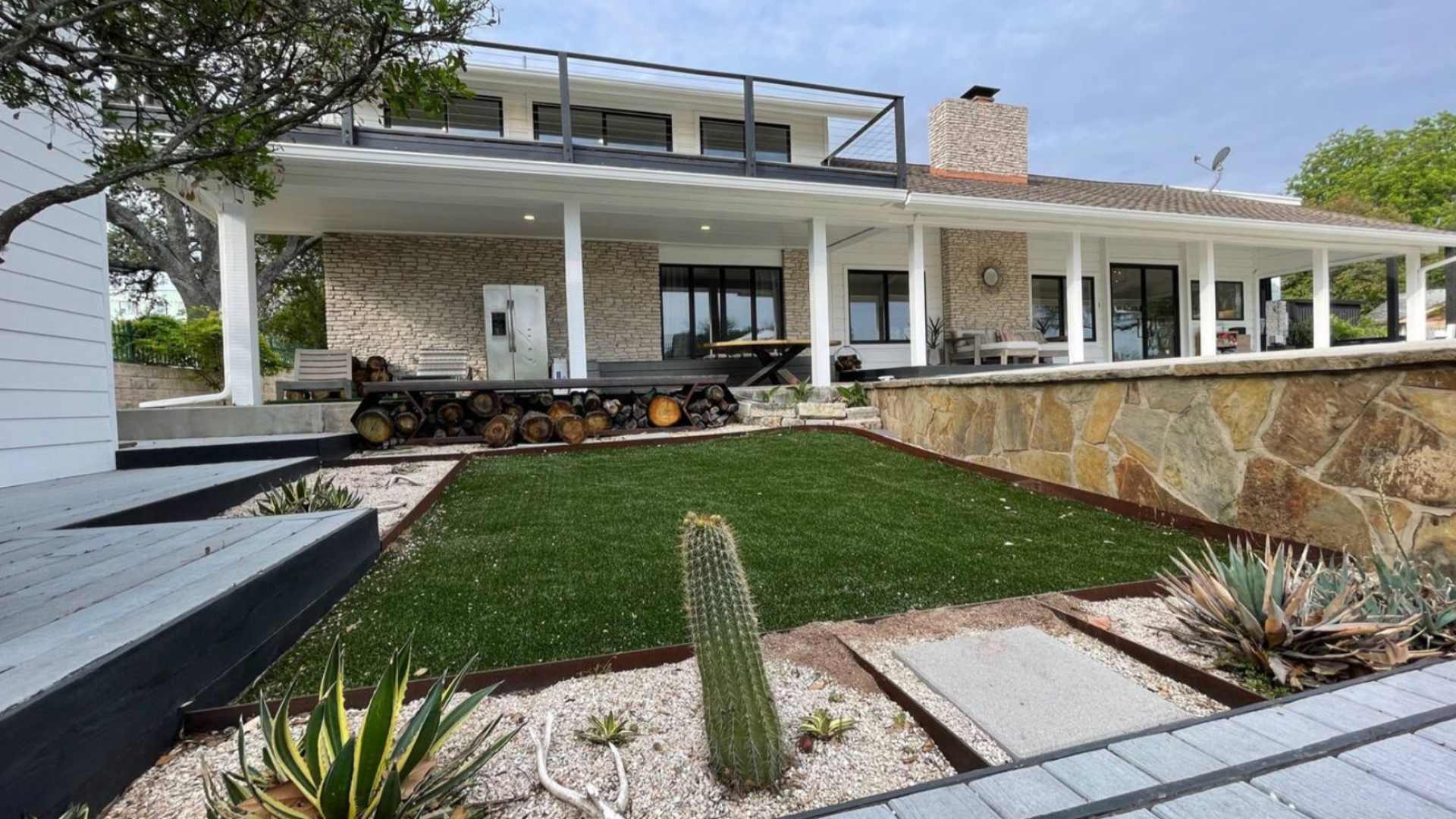 The back exterior of a modern house with stone and white siding, featuring a covered patio, a second-story balcony, and a landscaped yard with artificial turf, gravel, and desert plants.