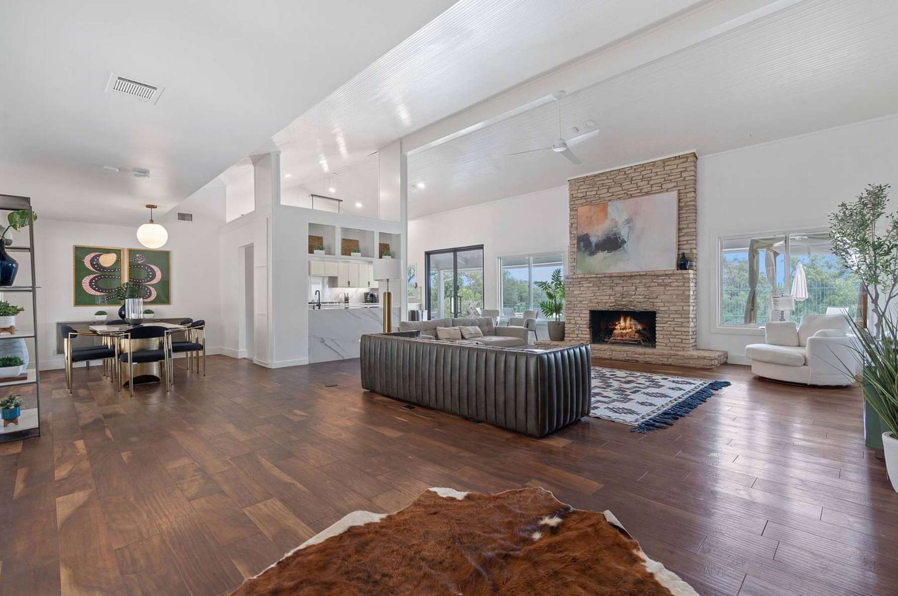 Expansive open-concept living space with dark hardwood floors, a large stone fireplace, and a comfortable seating arrangement, flowing into a modern dining area and a bright kitchen beyond.