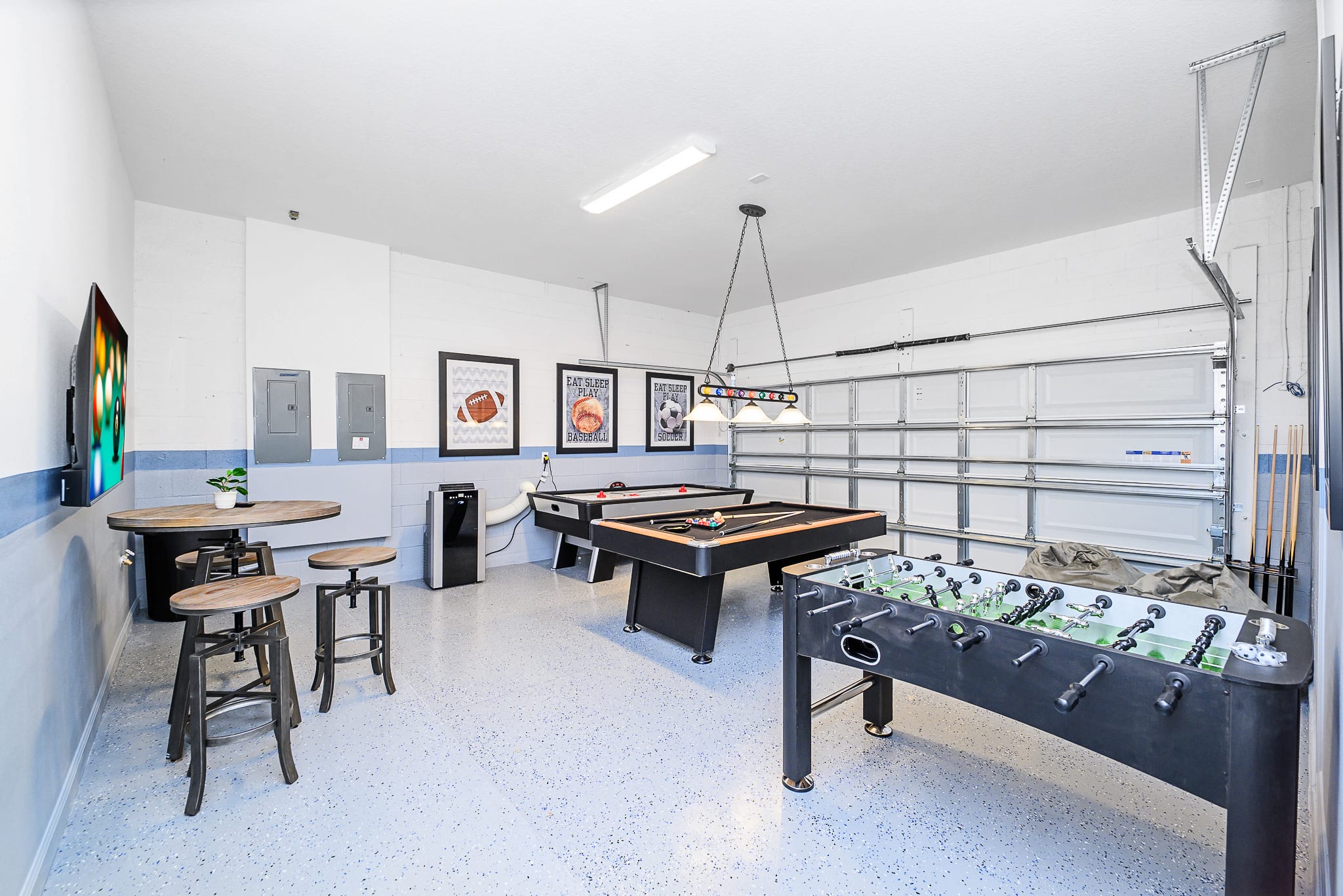 Another perspective of the bright game room reveals a foosball table, a pool table with an overhead light fixture, and an air hockey table. A high-top table with stools and a wall-mounted TV are also visible, all within the spacious, light-colored garage space.