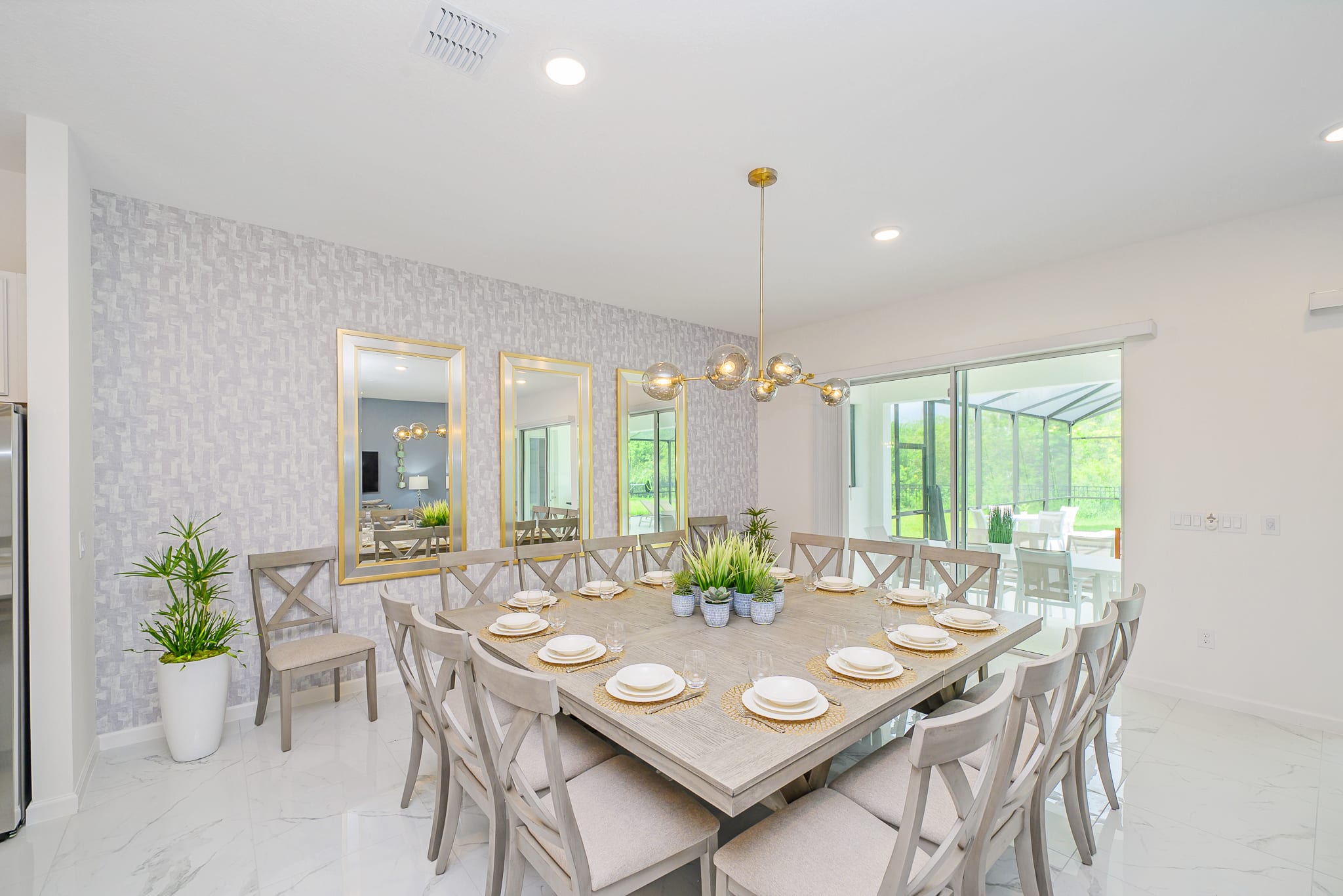 A large, square dining table with seating for many guests dominates this elegant dining room, featuring light grey wooden chairs and place settings. The room is adorned with patterned wallpaper and three large gold-framed mirrors, with a modern gold chandelier illuminating the space.