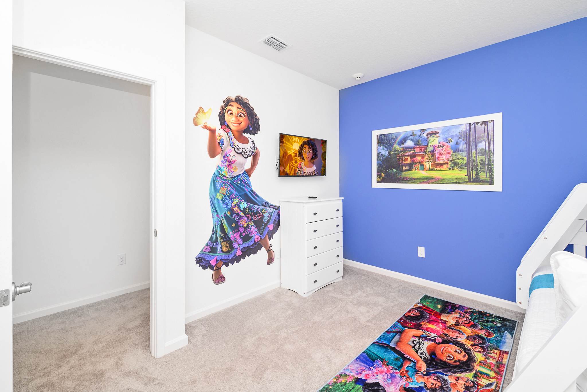 Another view of the Encanto-themed children's bedroom, showcasing a large Mirabel wall decal, a wall-mounted TV above a white dresser, and the blue accent wall with framed artwork.