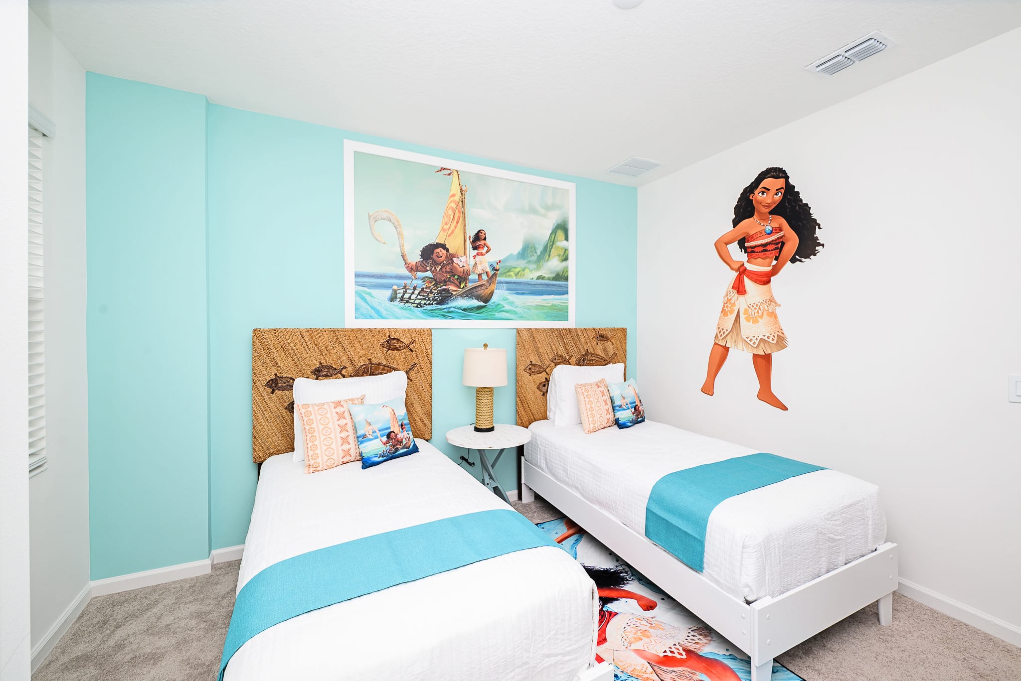 Vibrant kids' bedroom with a Moana theme, featuring two twin beds with woven headboards and blue accents. A large Moana mural and framed artwork decorate the light blue and white walls.