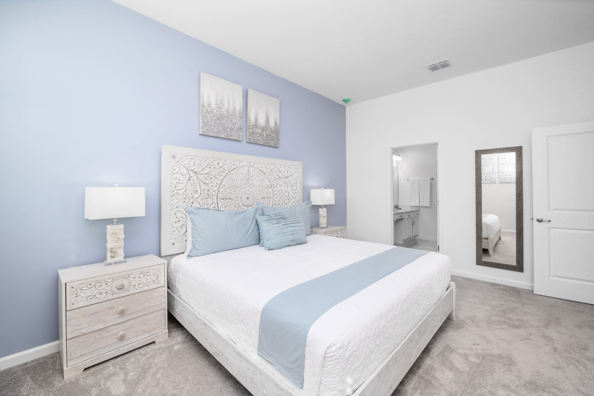 Inviting bedroom with a light blue accent wall and a white bed featuring a decorative headboard. A full-length mirror leans against the wall, and an open doorway leads into an en-suite bathroom.