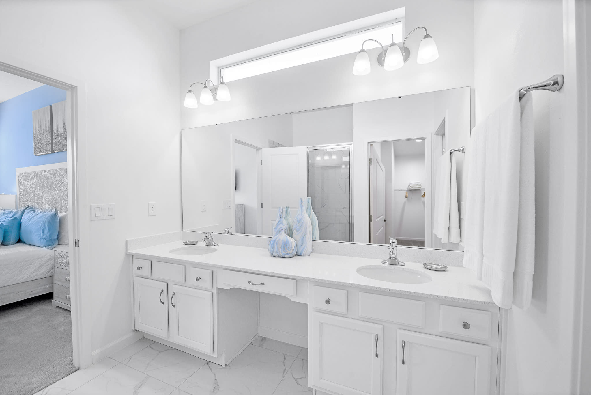 Spacious modern bathroom featuring a double vanity with white countertops, two sinks, and ample storage in white cabinets. A large mirror spans the width of the vanity, reflecting the bedroom beyond.