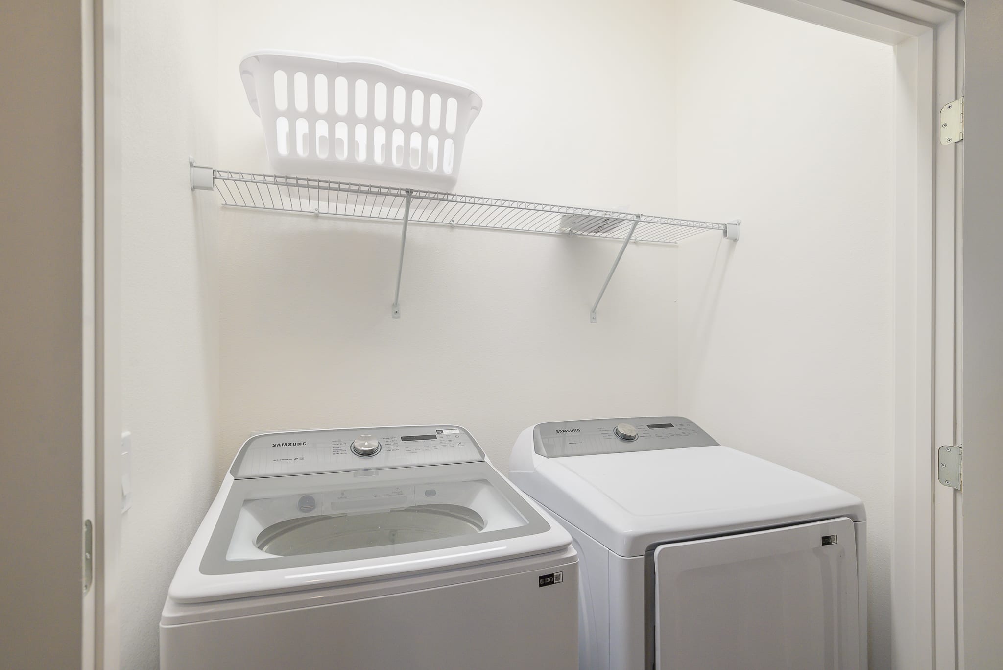 Compact laundry closet featuring a white top-load washing machine and a front-load dryer, with a wire shelf and a laundry basket mounted above.