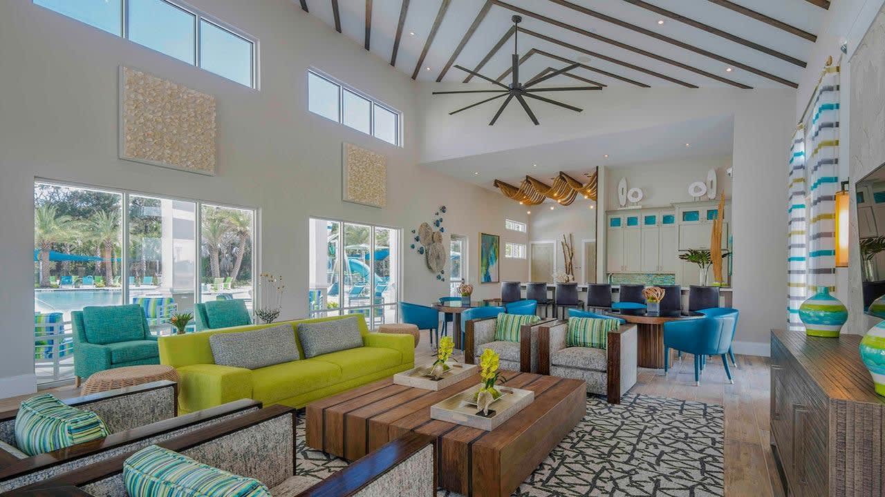 A spacious, modern communal lounge features high vaulted ceilings with exposed beams and a large decorative ceiling fan. Vibrant lime green and teal sofas and armchairs are arranged around wooden coffee tables, with a bar area visible in the background and large windows offering views of the outdoor pool.