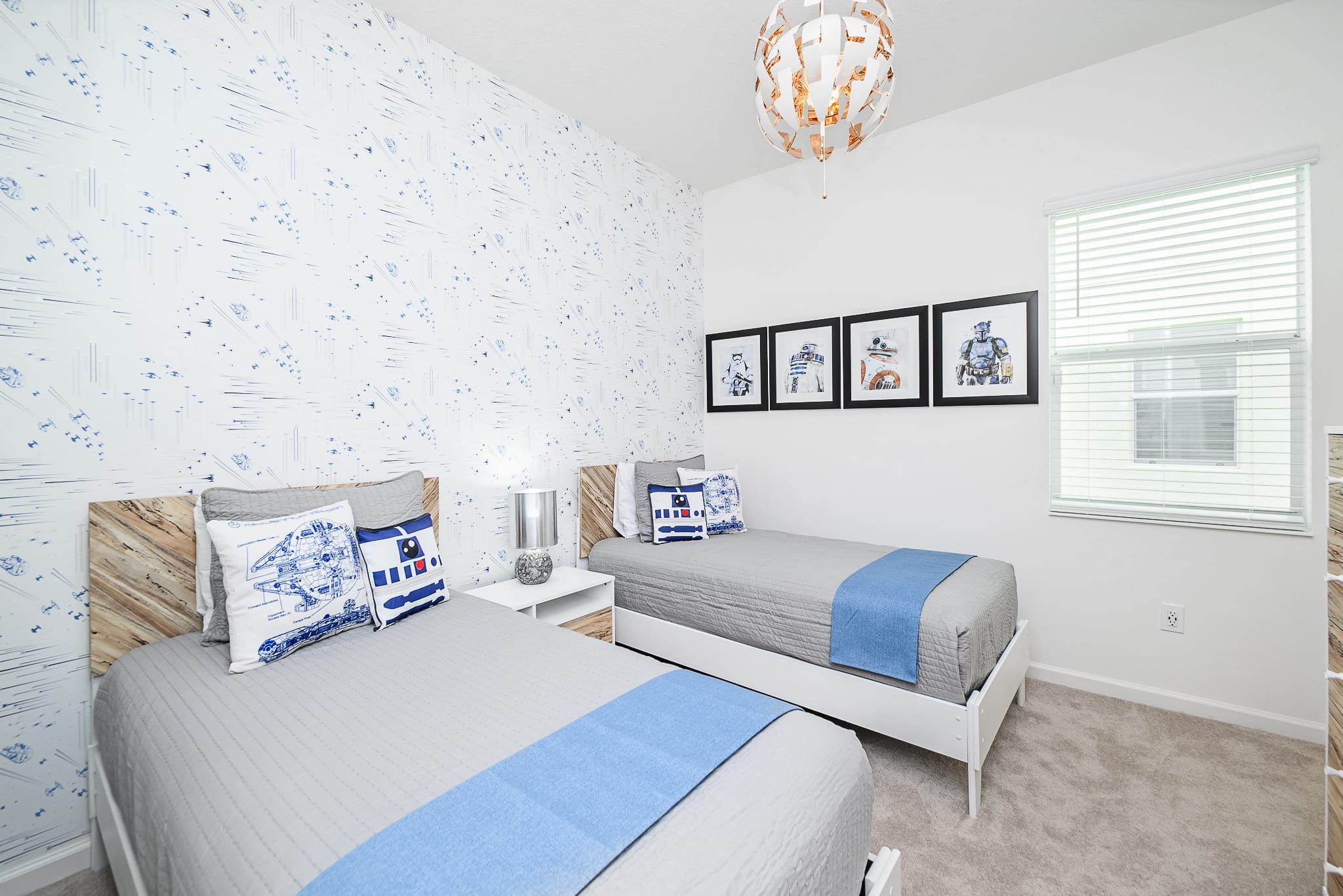 A Star Wars-themed bedroom featuring two twin beds with light grey bedding, blue accents, and character pillows, set against a white wall with blue spaceship wallpaper and framed Star Wars art.