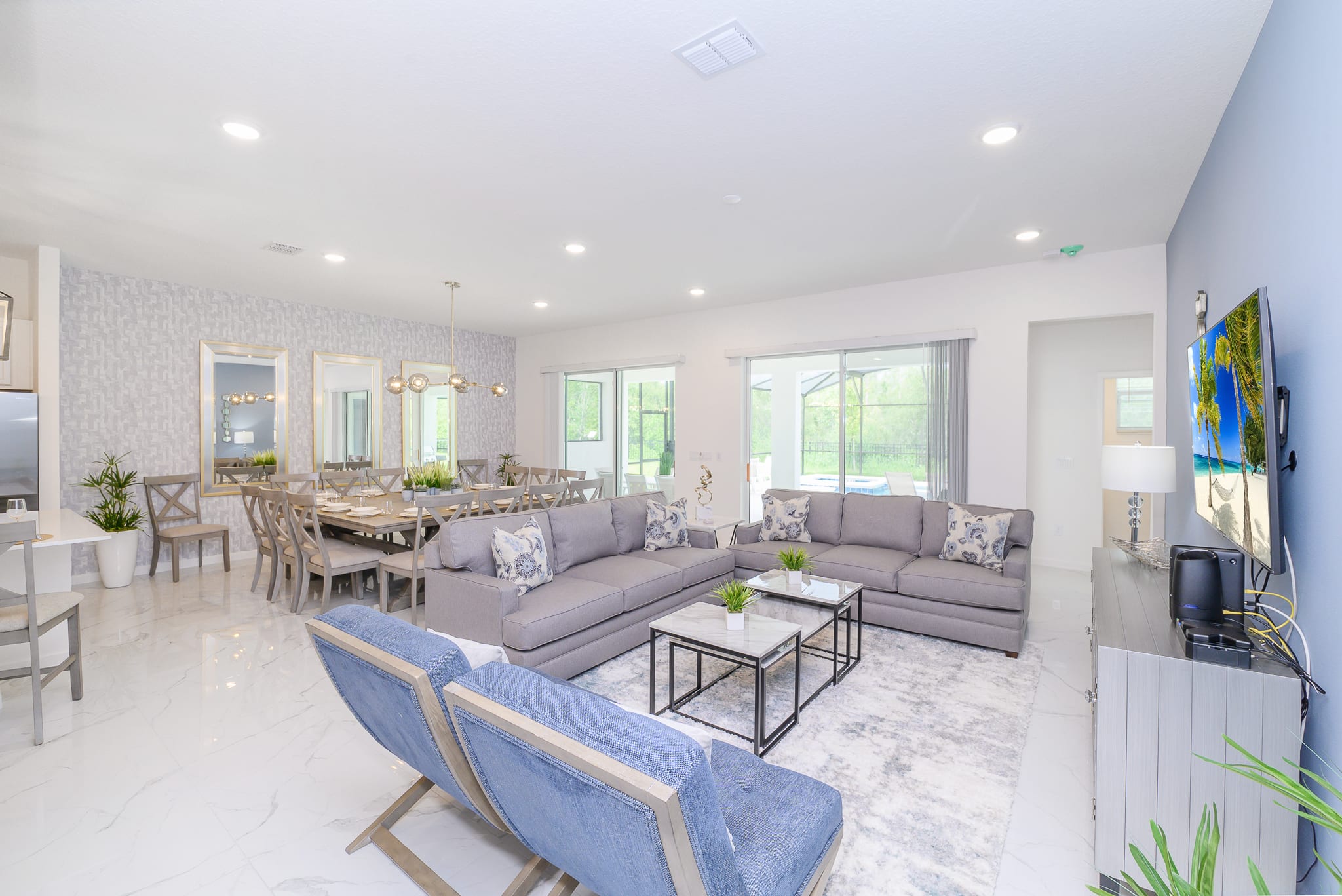 An open-concept living room features two grey sofas with patterned throw pillows, two modern coffee tables, and two blue accent chairs, all set on a light rug over white marble-look tile flooring. A large dining table with multiple chairs is visible in the background, along with sliding glass doors leading to an outdoor area.