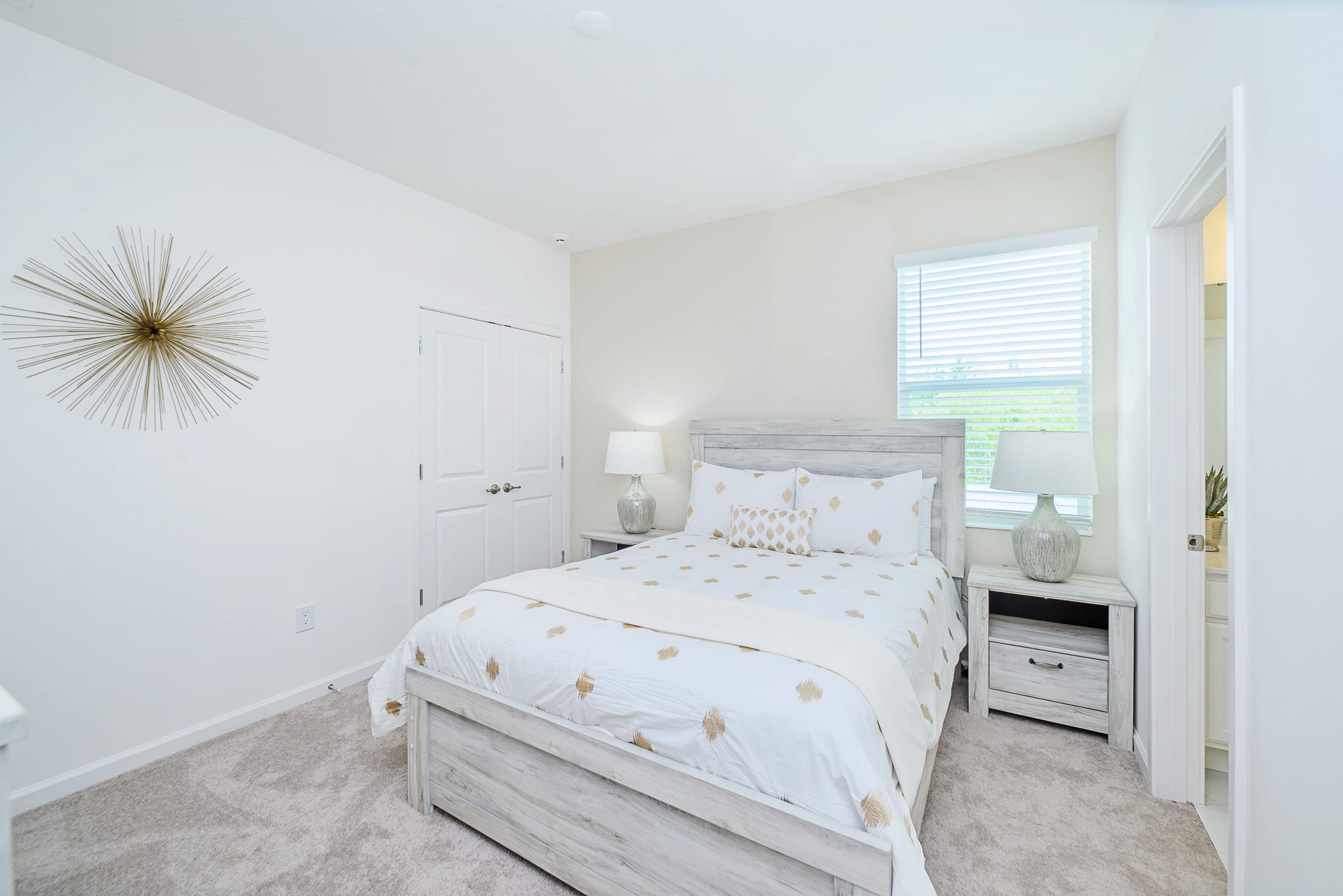 Cozy bedroom with a light wood-framed bed dressed in white and gold bedding, flanked by matching nightstands with lamps. A large gold starburst wall decor hangs above a closed closet.