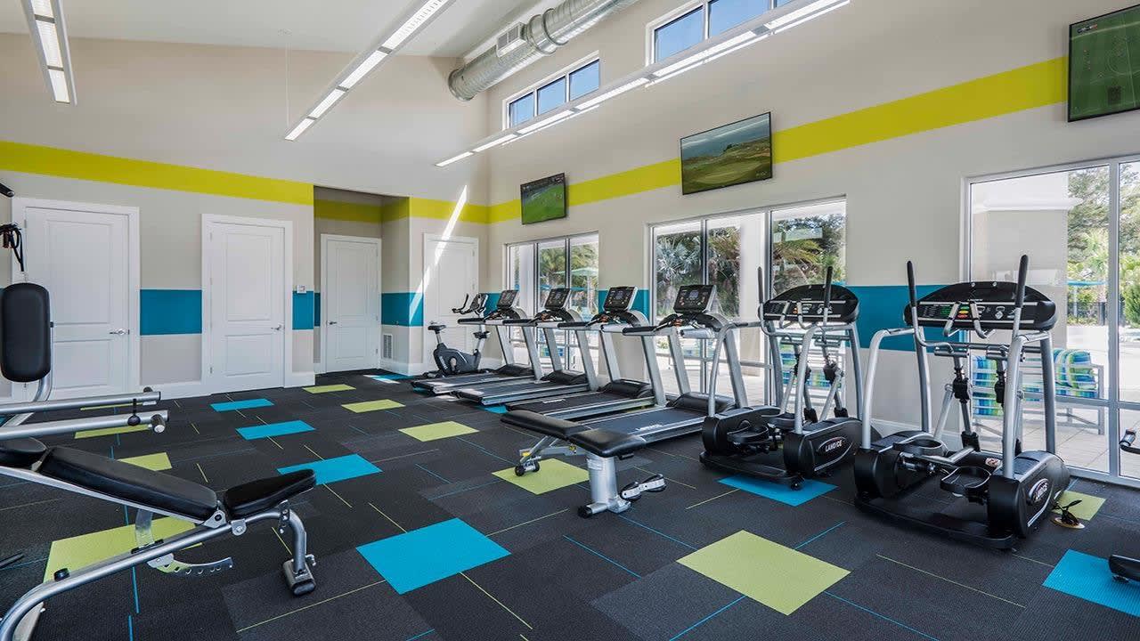 A bright, modern fitness center features a variety of exercise equipment including treadmills, ellipticals, and weight benches on a black, blue, and lime green patterned floor. Large windows overlook an outdoor area, and the walls are decorated with teal and yellow stripes.