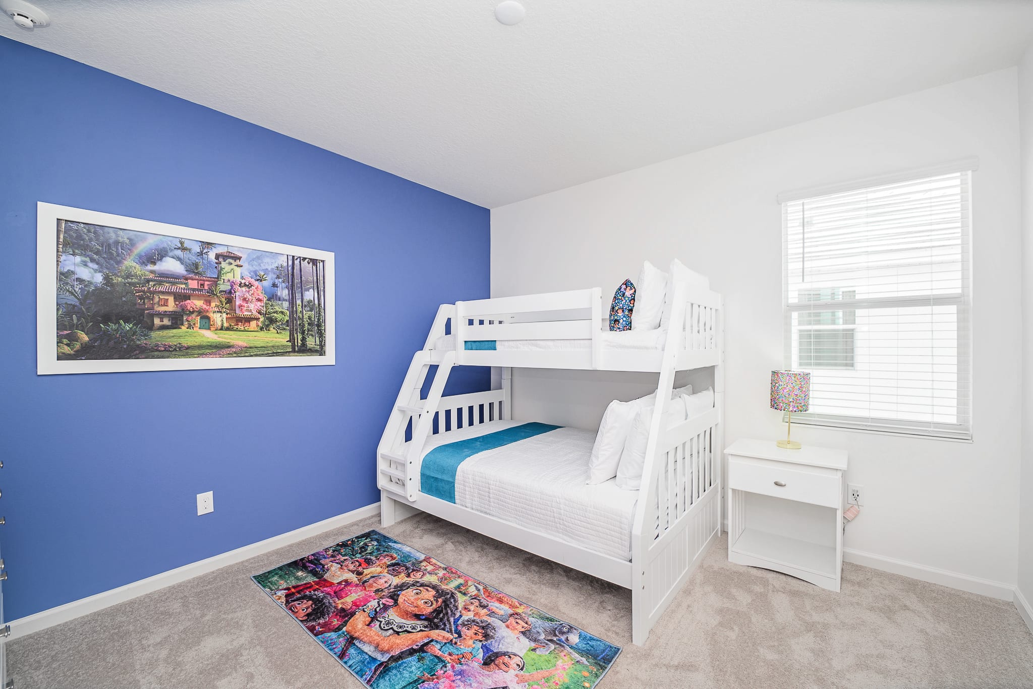 Vibrant children's bedroom featuring a white wooden bunk bed, a blue accent wall with a large framed Encanto artwork, and a colorful themed rug. A nightstand with a lamp is beside the bed.