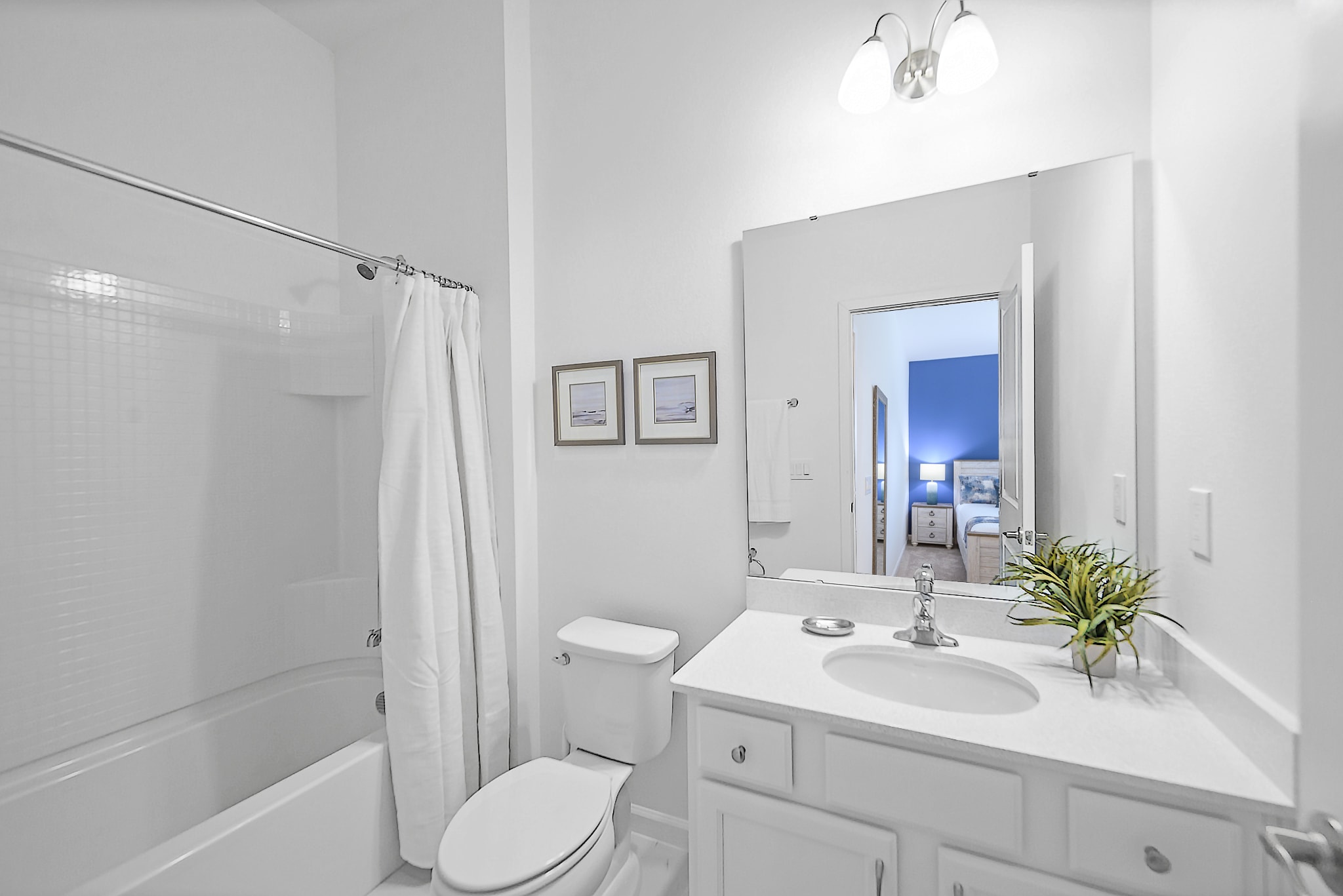 Clean white bathroom featuring a single vanity with a large mirror, a toilet, and a bathtub with a shower curtain. The mirror reflects a blue accent wall from an adjacent bedroom, adding a pop of color.
